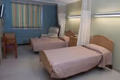 Two hospital-style beds in a shared nursing home room with privacy curtains, a recliner, bedside tables, and a wall-mounted TV.