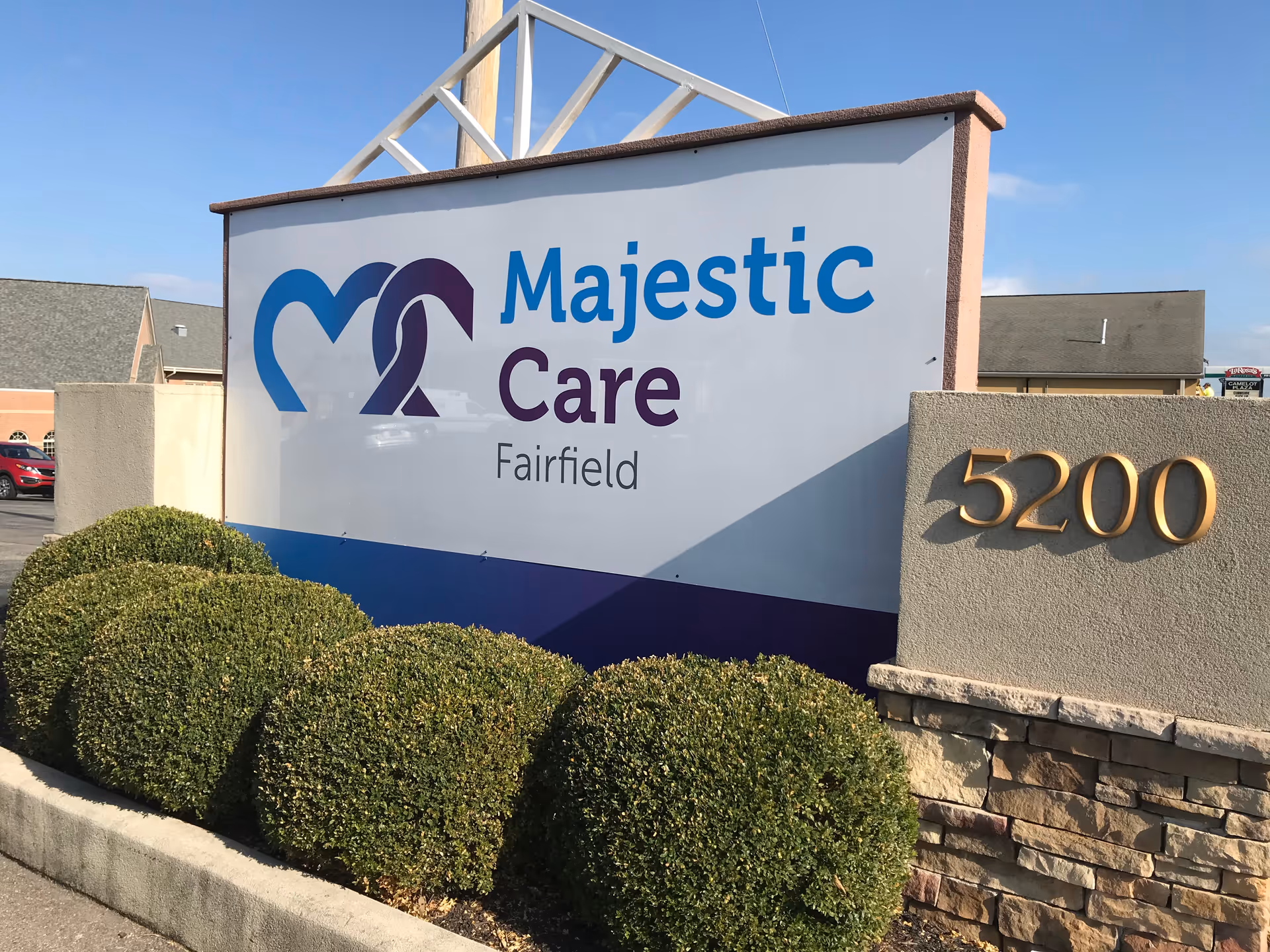 Outdoor sign for Majestic Care Fairfield with the address number 5200 displayed on a stone and stucco wall, surrounded by neatly trimmed bushes under a clear blue sky.
