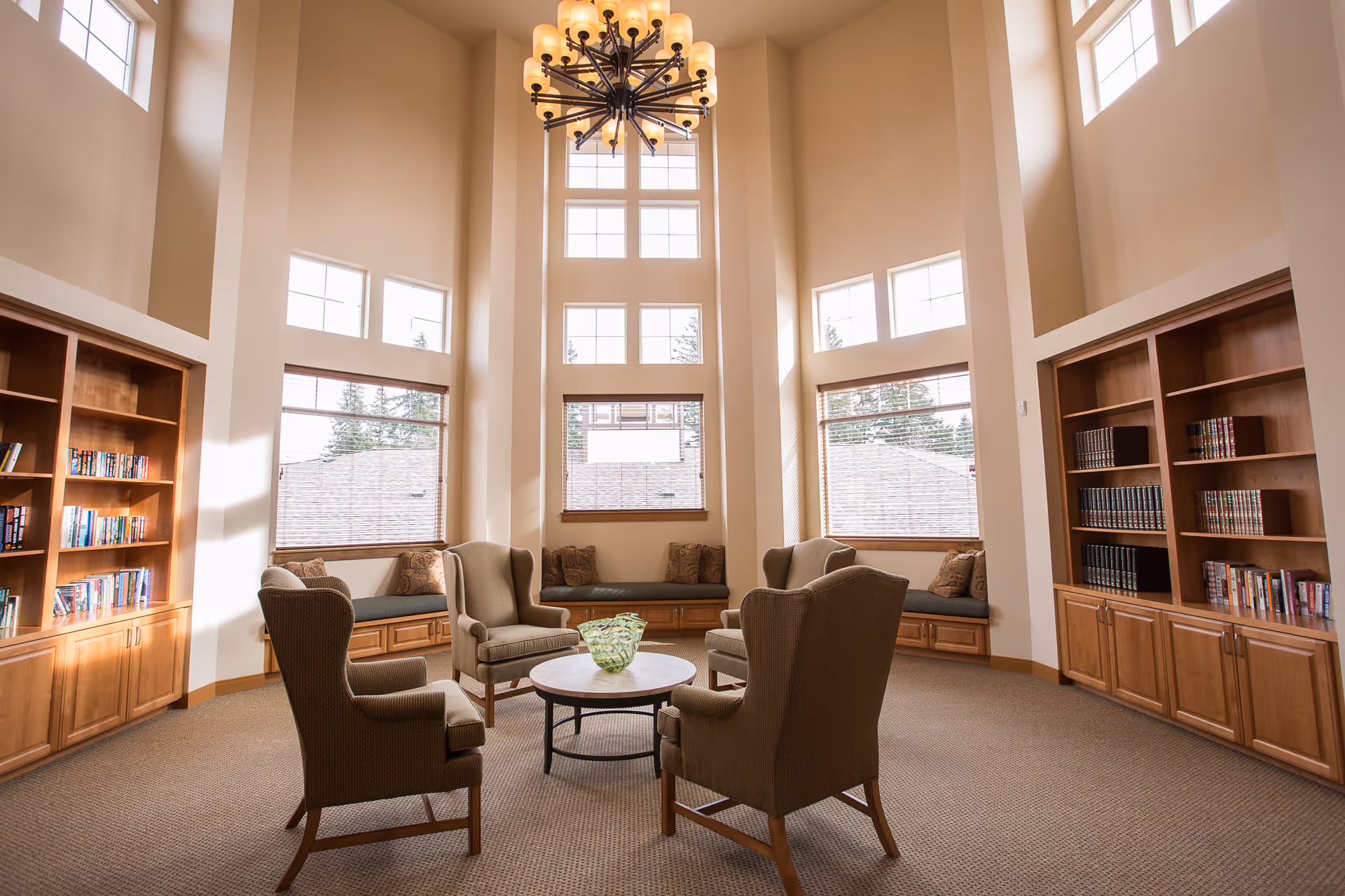 A bright and spacious living room area with high ceilings and large windows letting in natural light. The room features four upholstered armchairs arranged around a round coffee table with a decorative glass bowl. Built-in wooden bookshelves filled with books line the walls on either side, and cushioned window seats are beneath the windows.
