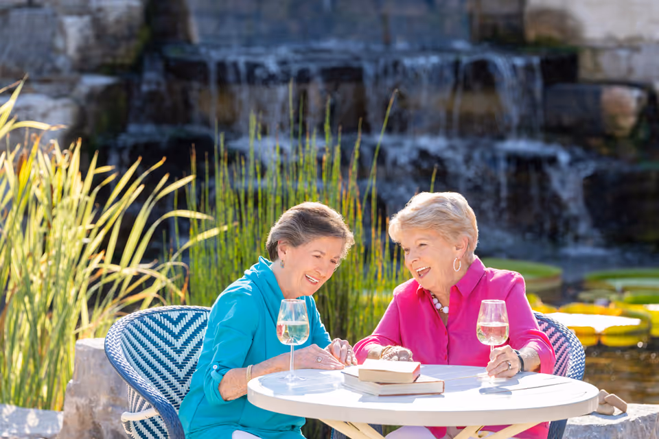 Two elderly women sitting at a round white table outdoors, enjoying drinks and smiling while talking. Behind them is a small waterfall feature surrounded by greenery and water plants.