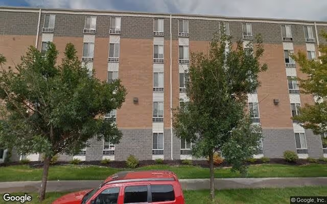 Exterior view of a multi-story brick and concrete building with several windows, surrounded by green trees and grass. A red vehicle is parked in front of the building under a cloudy sky.