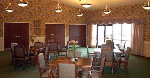 A dining room with several wooden tables and chairs arranged neatly. The room has floral wallpaper, green carpet, and large windows with curtains allowing natural light to enter. There are decorative light fixtures hanging from the ceiling and some floral arrangements on the tables.