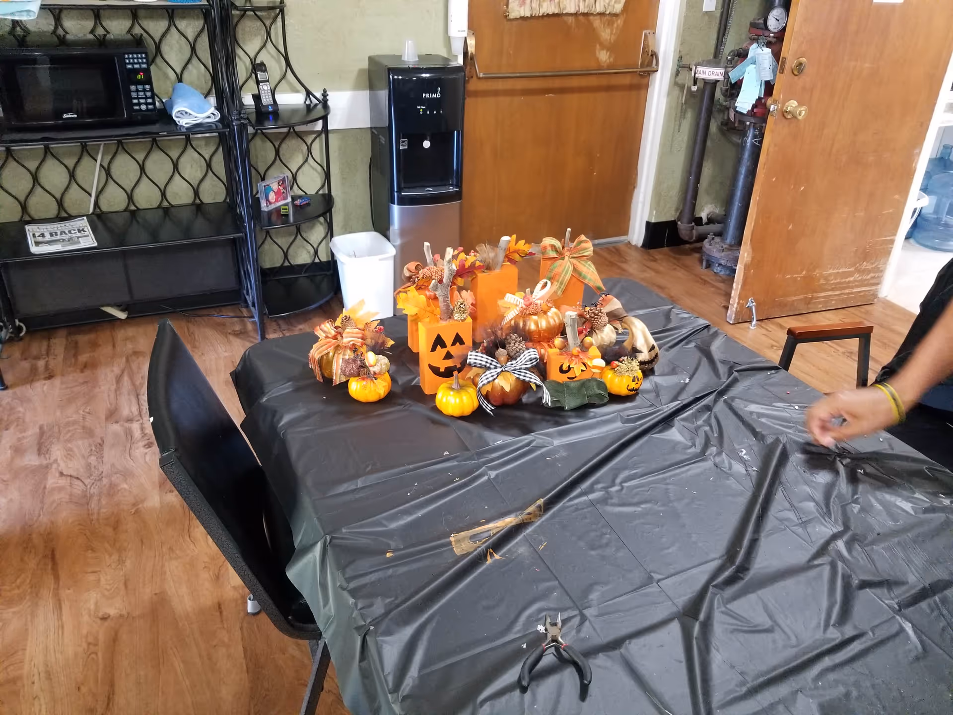A room with a table covered in a black plastic tablecloth, decorated with small Halloween-themed pumpkins and fall decorations. There is a black chair next to the table, a water dispenser against the wall, a microwave on a metal shelf, and a wooden door in the background. A person's arm is visible on the right side of the image.