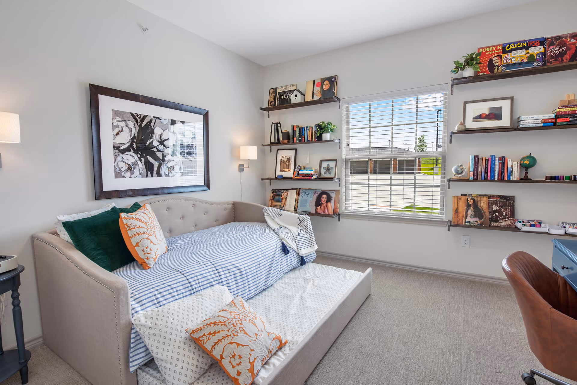 Bright bedroom with a daybed, decorative pillows, wall art, and shelving by a window.