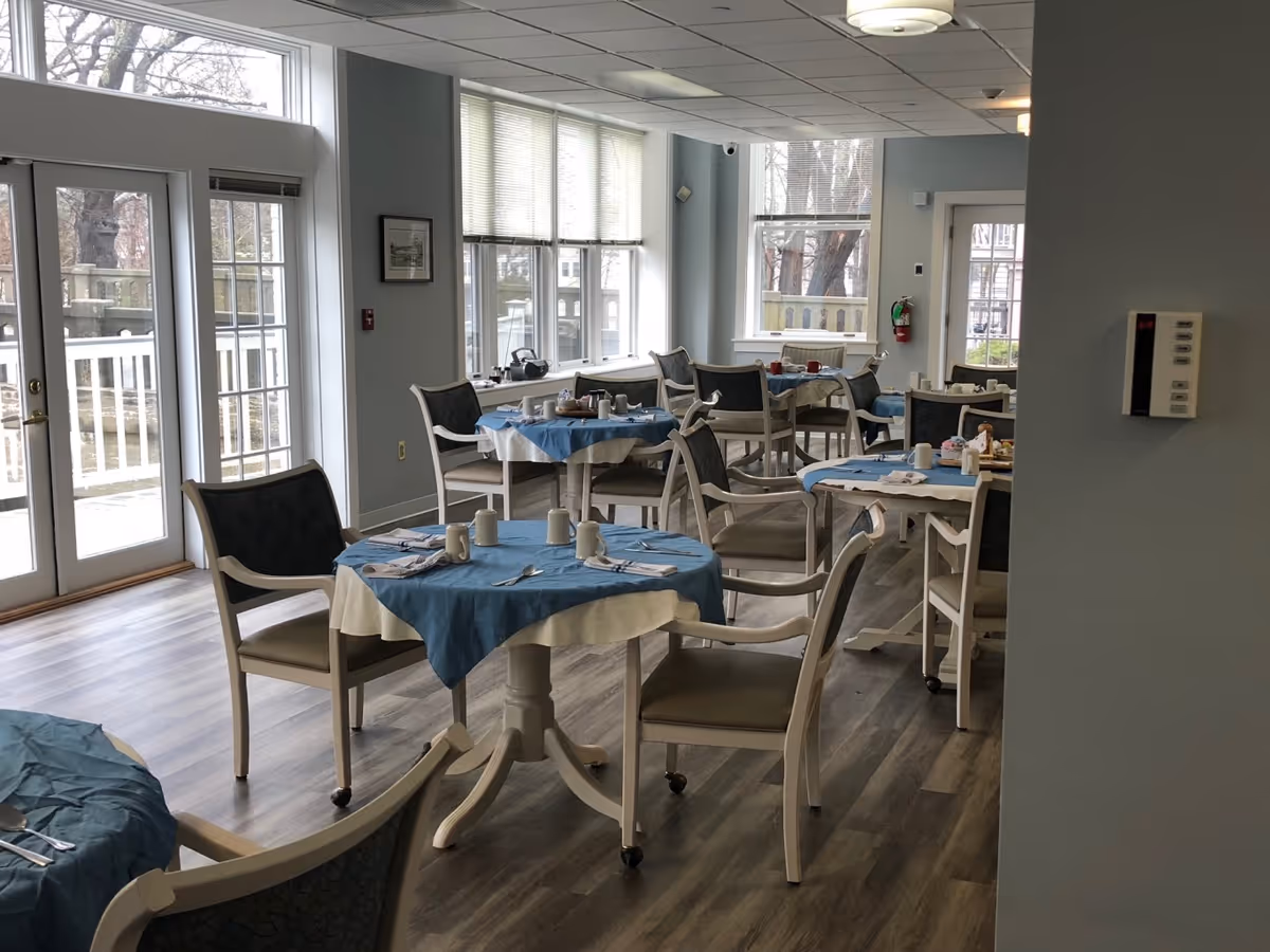 A bright dining room with several round tables covered with blue and white tablecloths. Each table is set with white mugs, napkins, and silverware. The room has large windows letting in natural light and light-colored wooden flooring. Chairs with cushioned seats and backs surround the tables. There is a door leading outside and a thermostat on the wall.