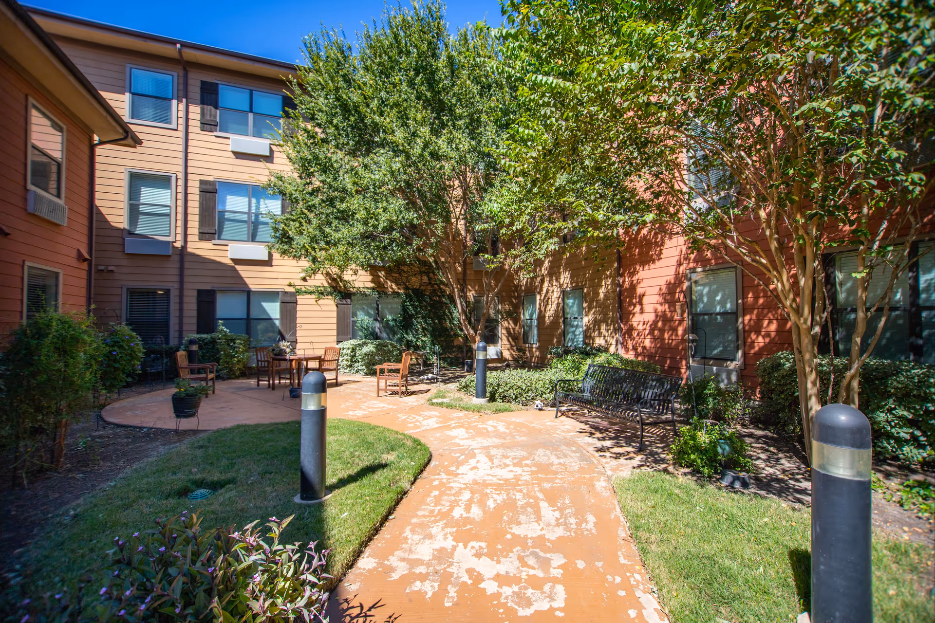 Outdoor courtyard area at Isle at Watercrest Bryan with a paved walking path, green grass, trees, shrubs, outdoor seating including chairs and a bench, and a multi-story building with windows surrounding the courtyard.