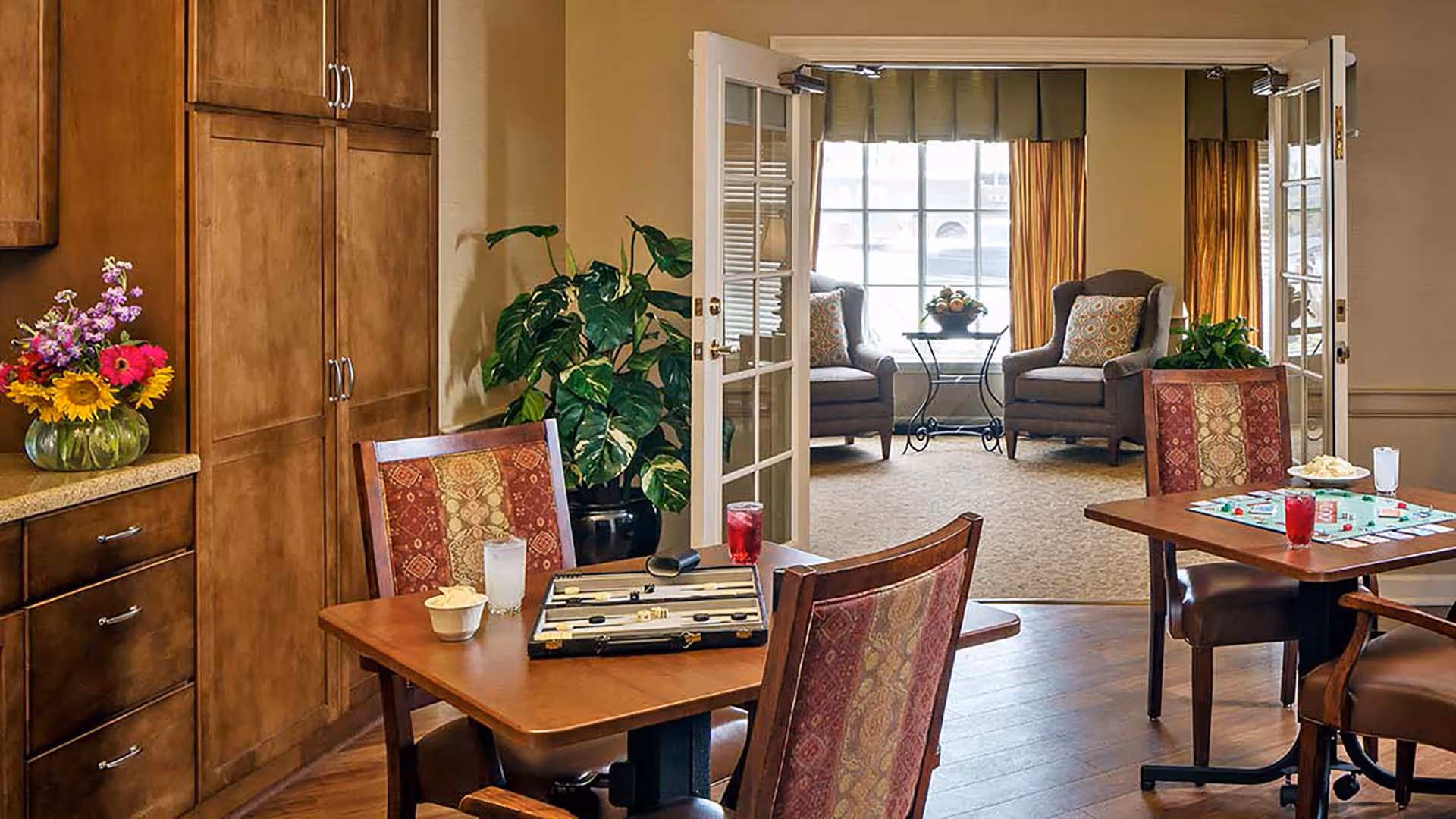 A cozy interior space featuring two wooden tables with patterned chairs. On the tables are board games, drinks, and snacks. To the left, there are wooden cabinets with a vase of colorful flowers on the countertop. In the background, French doors open to a sitting area with two armchairs, a small table with a floral arrangement, and large windows with curtains.