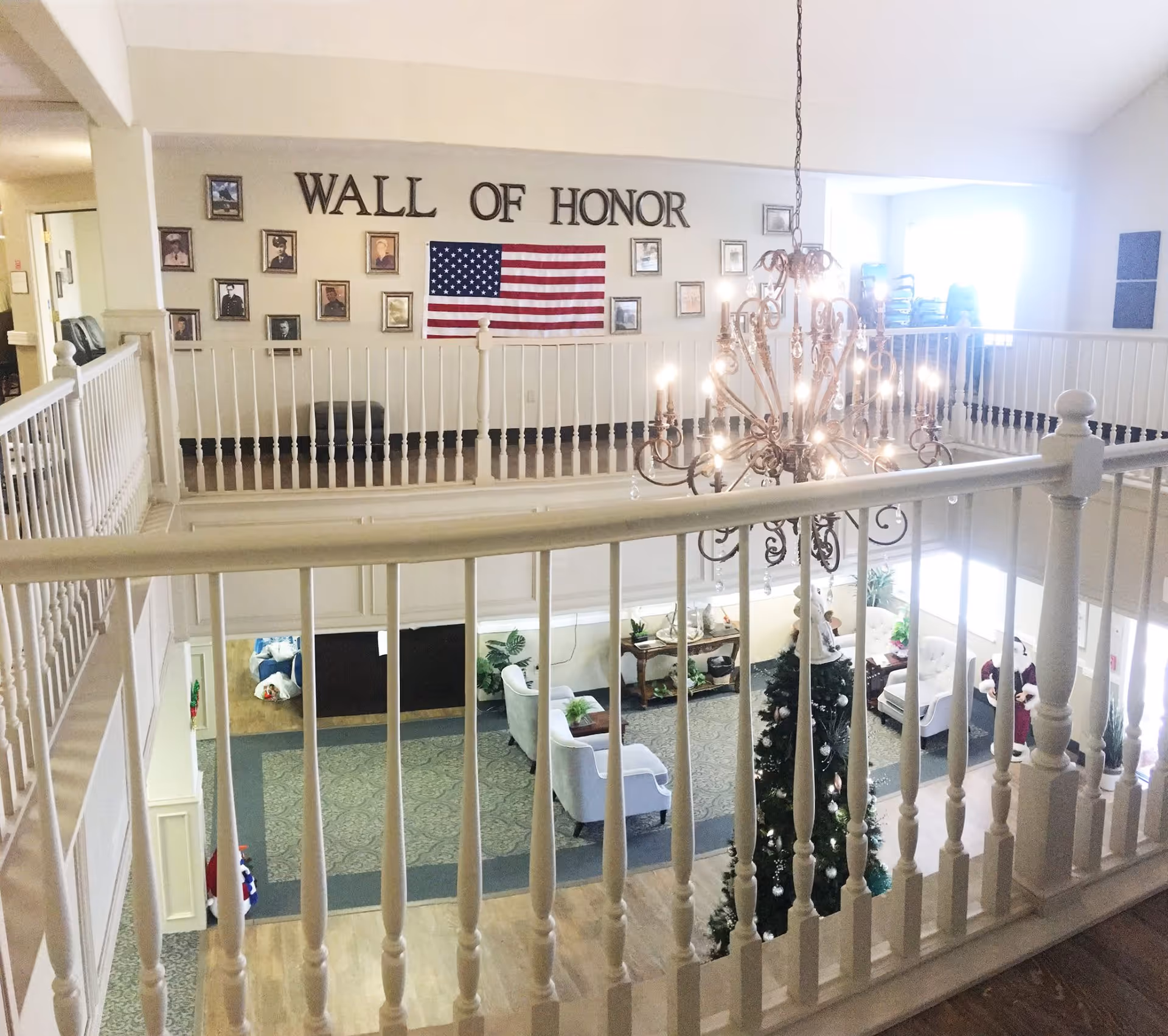 Interior view of a senior living facility showing a two-story common area with white railings, a chandelier, and a decorated Christmas tree. On the upper wall, there is a 'Wall of Honor' display with framed photos and an American flag. The lower level has seating areas with armchairs and a carpeted floor.