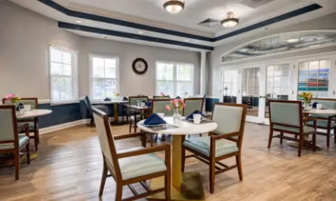 A bright dining room with several round tables and wooden chairs with light cushions. Each table is set with napkins and small flower arrangements. Large windows let in natural light, and a clock is mounted on the wall. The room has a wooden floor and a ceiling with recessed lighting and decorative molding.