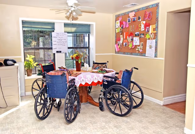 A small dining area in an assisted living facility with a round table covered by a lace tablecloth and a red underlay. Four wheelchairs are positioned around the table. The room has a bulletin board on the wall decorated with colorful papers and photos. A window with blinds lets in natural light, and there are plants on the windowsill. The floor has a light-colored patterned linoleum.