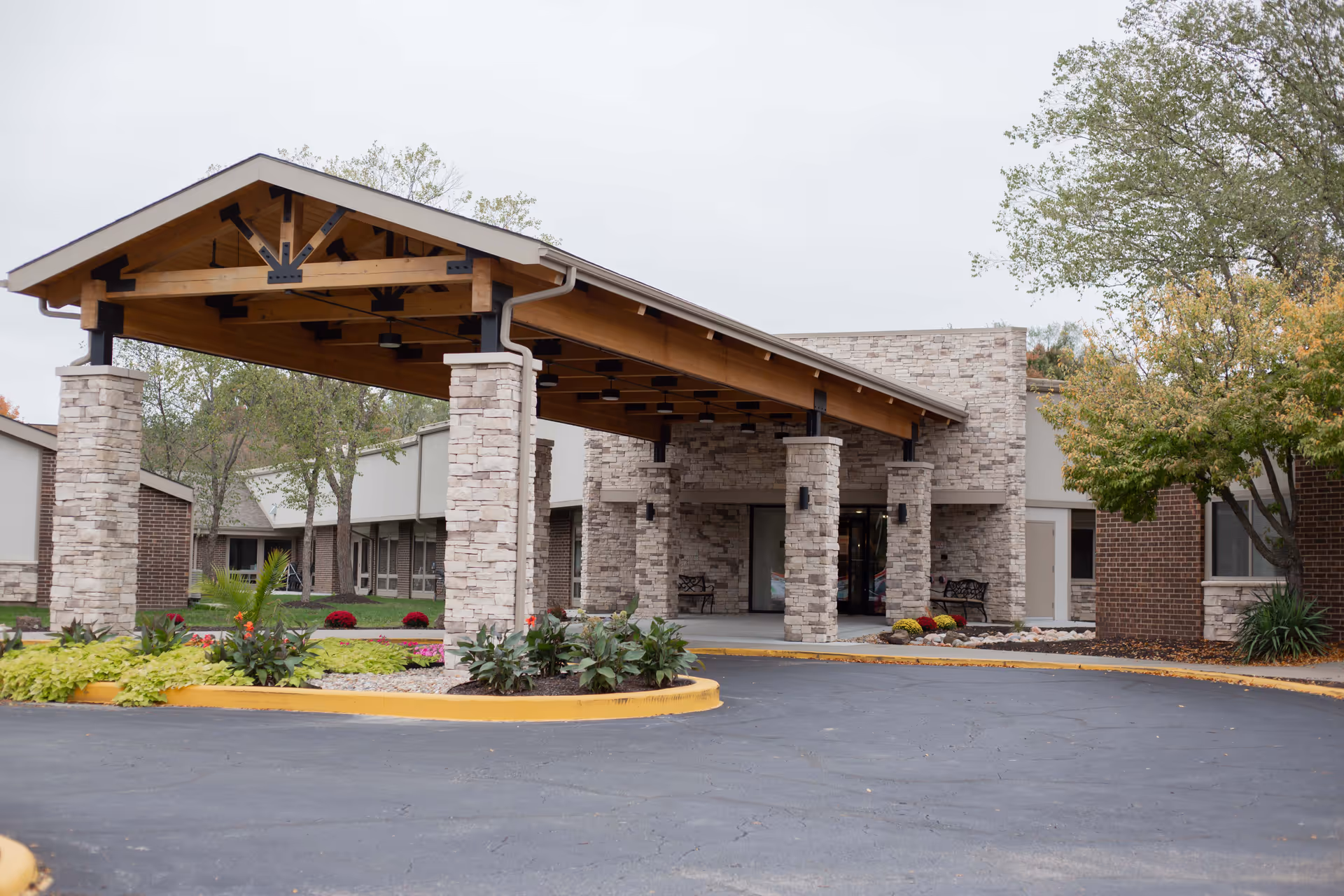 Entrance of The Grand Royale Senior Living facility featuring a covered driveway with stone pillars and wooden beams, surrounded by landscaped greenery and trees.