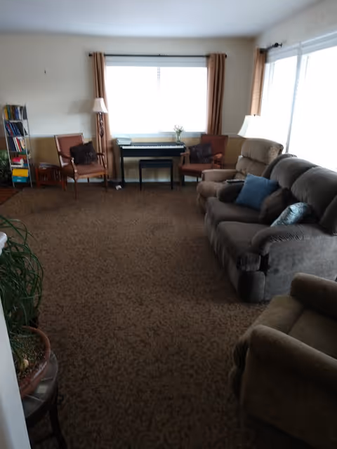 A cozy living room with brown carpet and beige walls. The room features a dark brown sofa with blue and brown cushions, two armchairs, a small table with a lamp, a keyboard piano under a window with beige curtains, and a bookshelf filled with books in the corner.