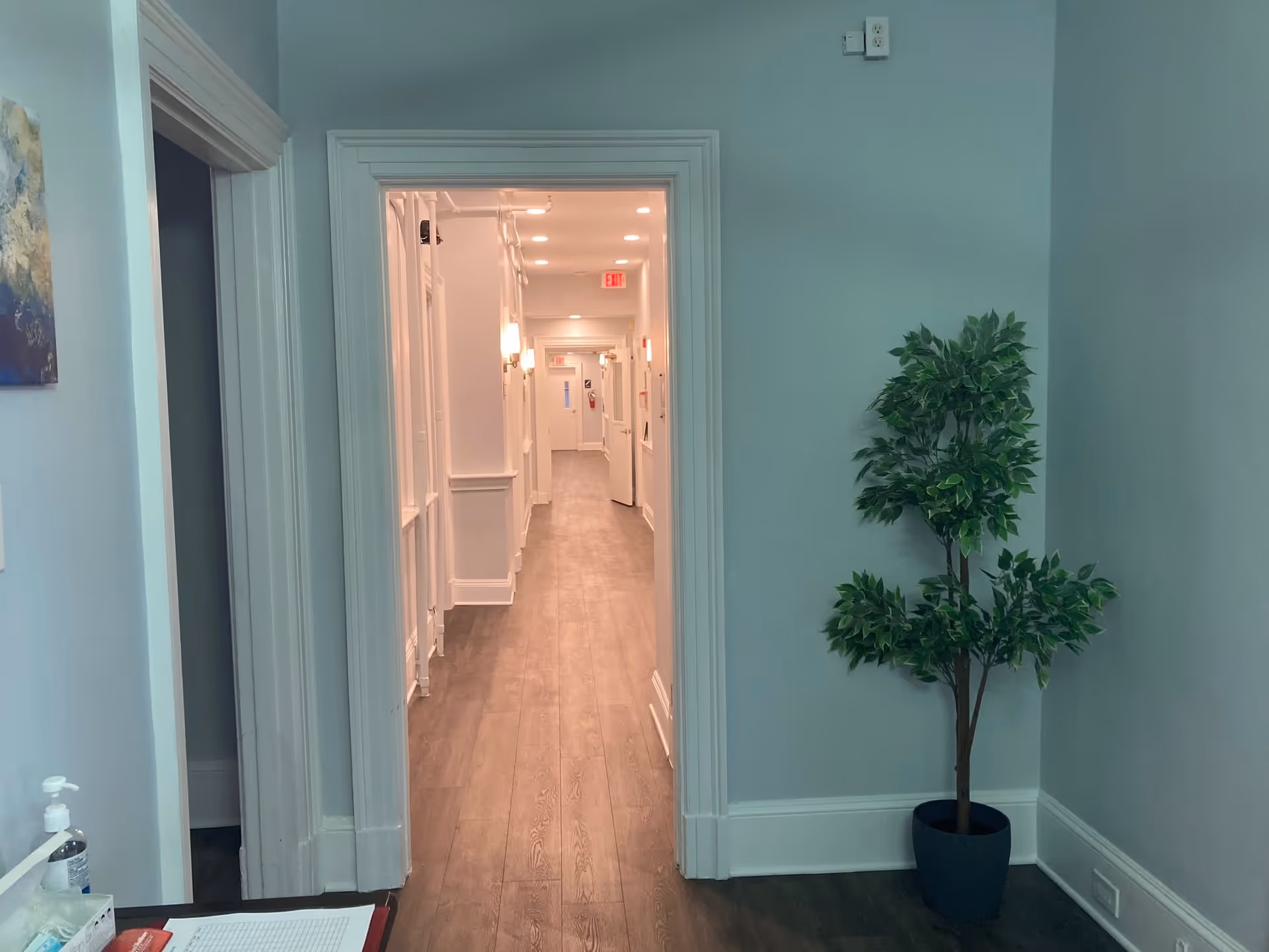 View of a hallway in a senior living community with wooden flooring, light blue walls, white door frames, and a potted plant on the right side. The hallway is well-lit with ceiling lights and wall sconces, and there is an exit sign visible at the end.