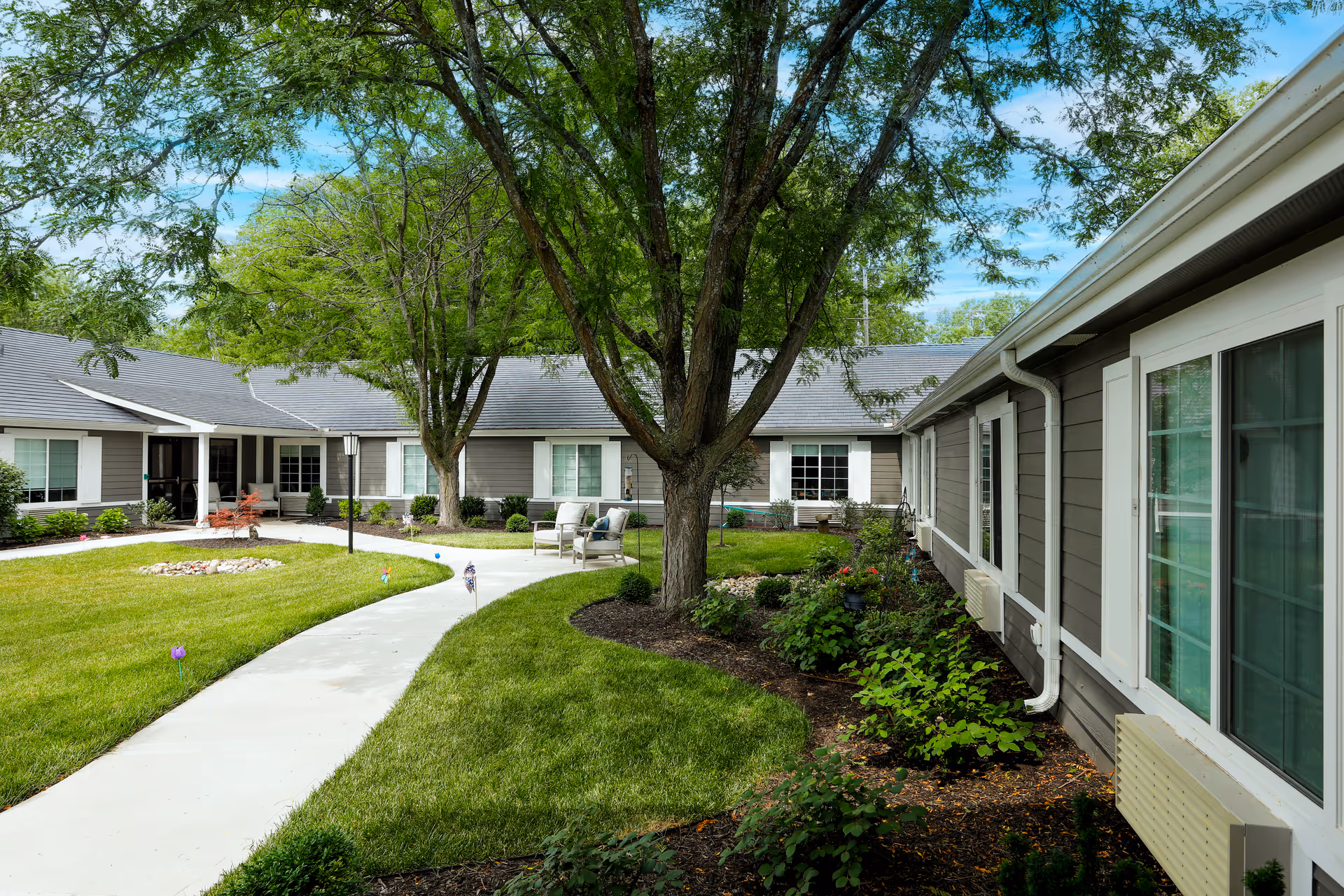 Outdoor courtyard area of Homestead Estates Assisted Living of Leawood featuring a curved concrete walkway, green grass, trees, shrubs, and seating areas with chairs. The building surrounds the courtyard with windows and a gray exterior.