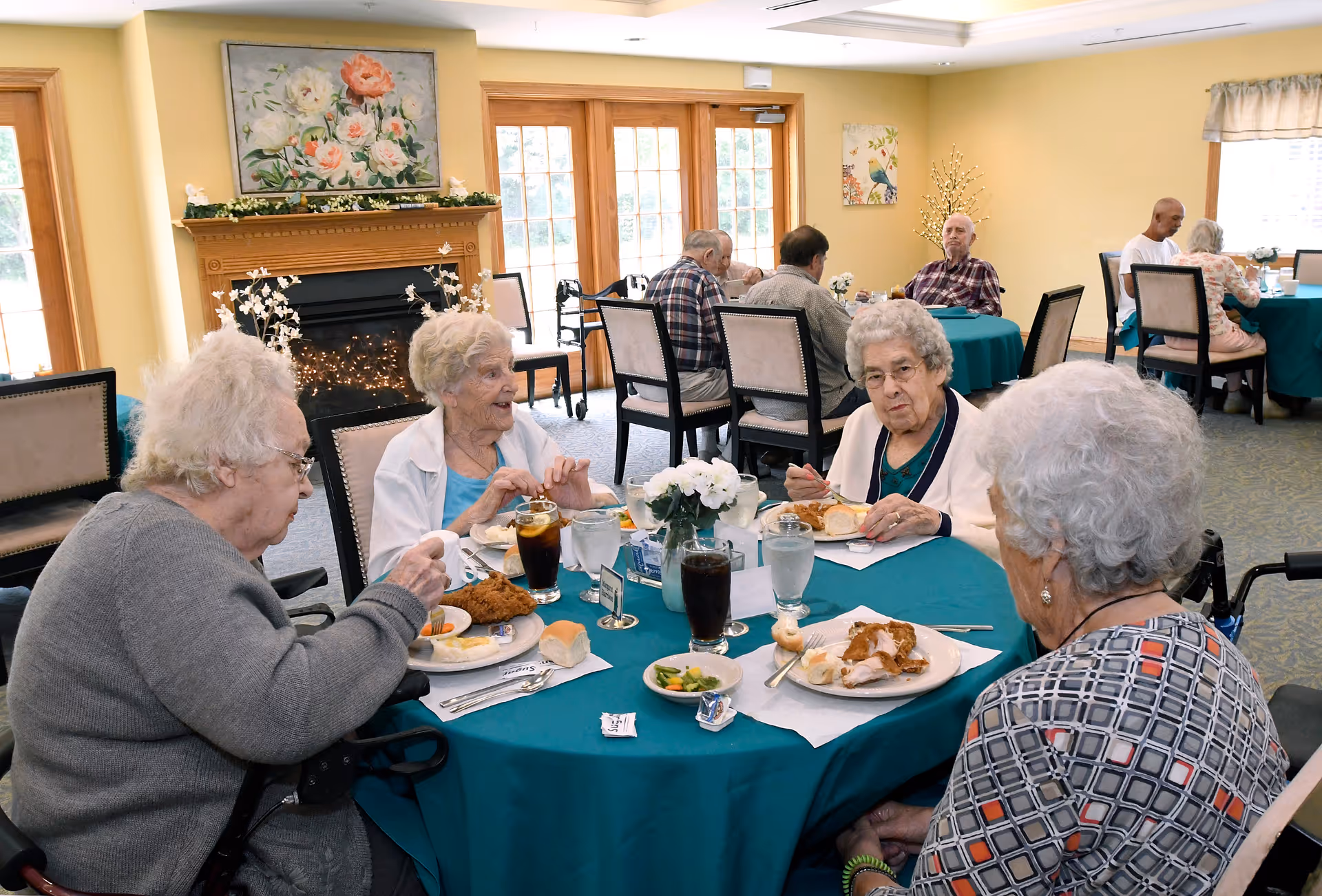 A group of elderly people sitting around a round table with a teal tablecloth, eating a meal together in a dining room. The room has large windows, a fireplace with floral artwork above it, and other tables with people dining in the background.