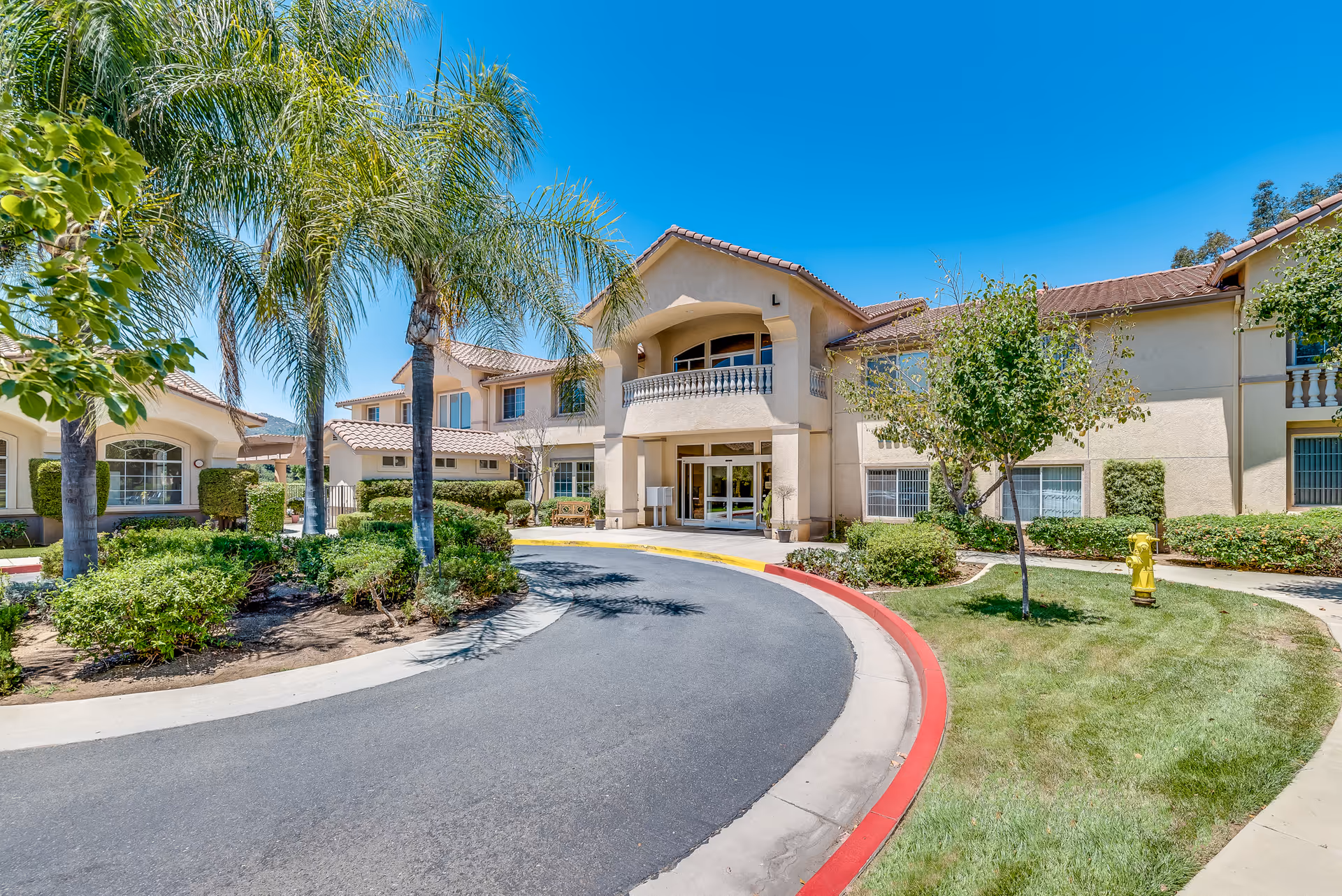 Two-story stucco senior living building with a covered main entrance, circular driveway, palm trees, and landscaped grounds under a clear blue sky.