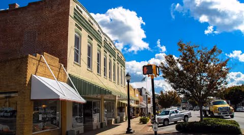 Street view of a small town with brick and painted buildings, parked cars, a tree, and a blue sky with scattered clouds.