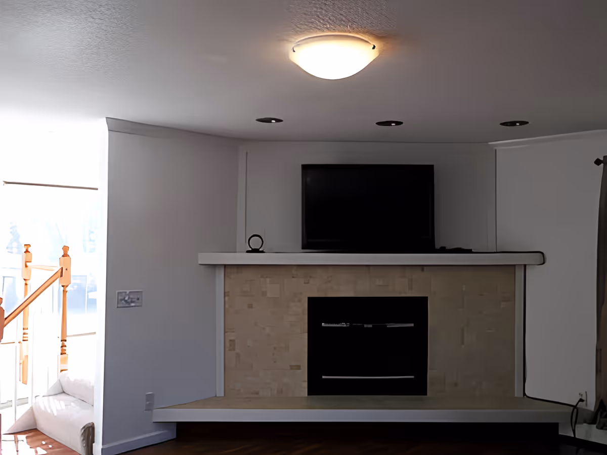 A living room area featuring a modern fireplace with a beige tiled surround and a flat-screen TV mounted above it. The walls are painted white, and there is a ceiling light fixture above. To the left, there is a staircase with wooden handrails and a large window letting in natural light. A curtain is partially visible on the right side.