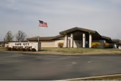 Single-story nursing home building with an American flag on a pole and a stone sign reading "Cave City Nursing Home" in the front lawn.