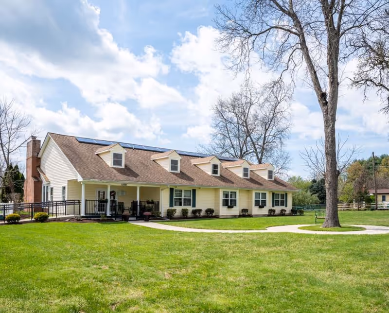 A single-story yellow building with a brown roof and several dormer windows, surrounded by a well-maintained green lawn and a few leafless trees under a partly cloudy sky.