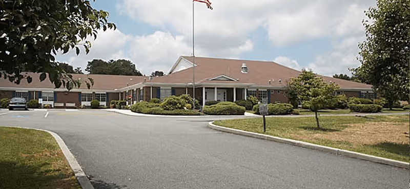 Exterior view of a single-story brick building with a red roof, surrounded by greenery and a paved driveway leading to the entrance under a partly cloudy sky.