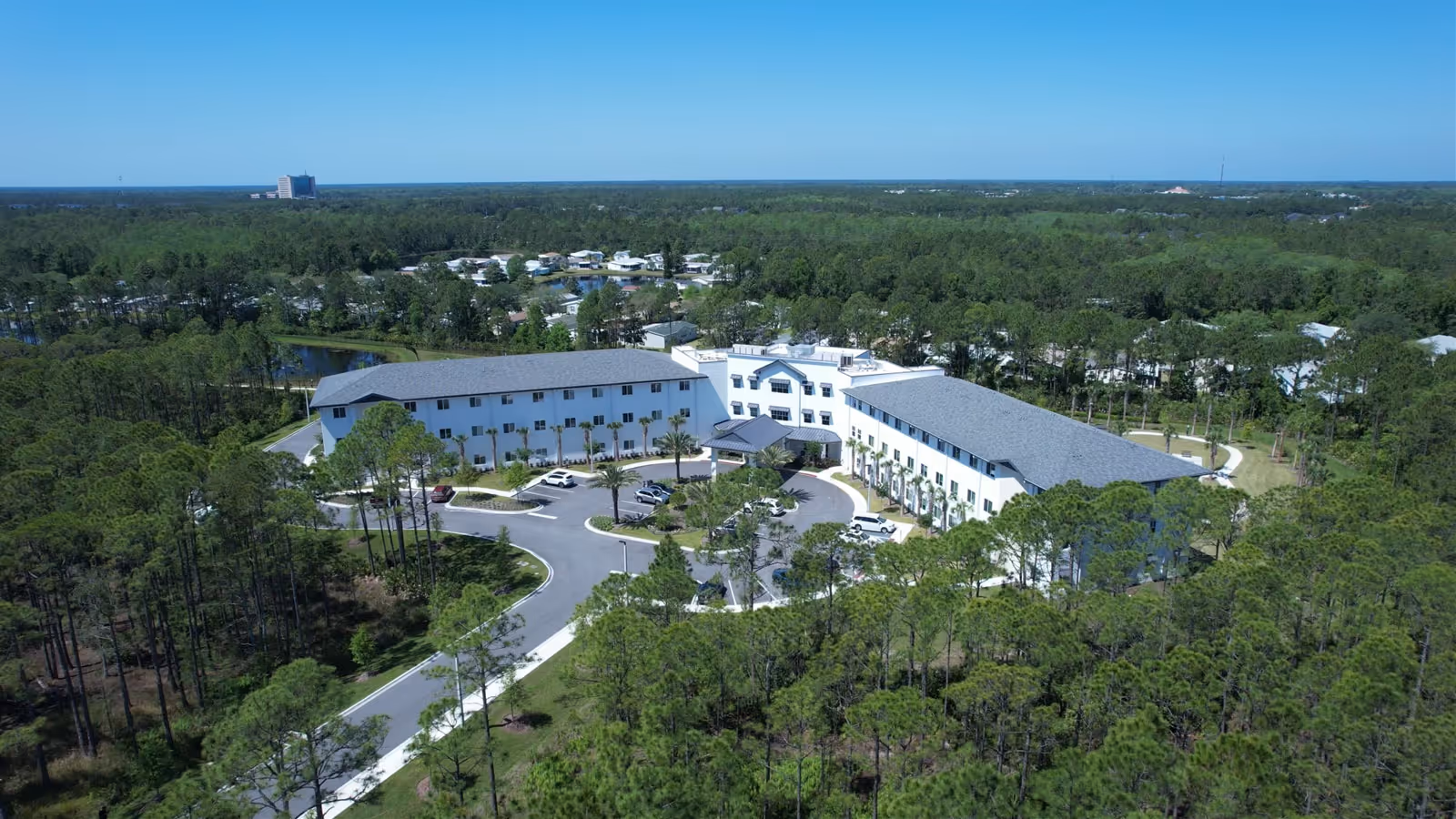 Aerial view of a large, white, three-story assisted living facility surrounded by dense green trees and forest. The building has a U-shaped layout with a circular driveway and parking area in front. The sky is clear and blue.