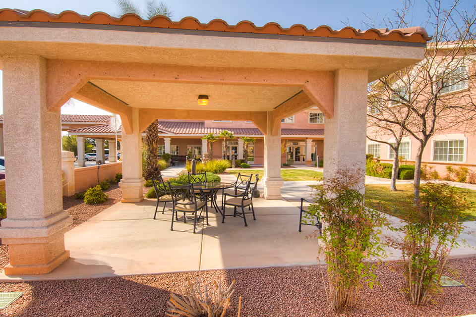 Covered outdoor patio with a metal table and chairs in a landscaped courtyard in front of a senior living building.