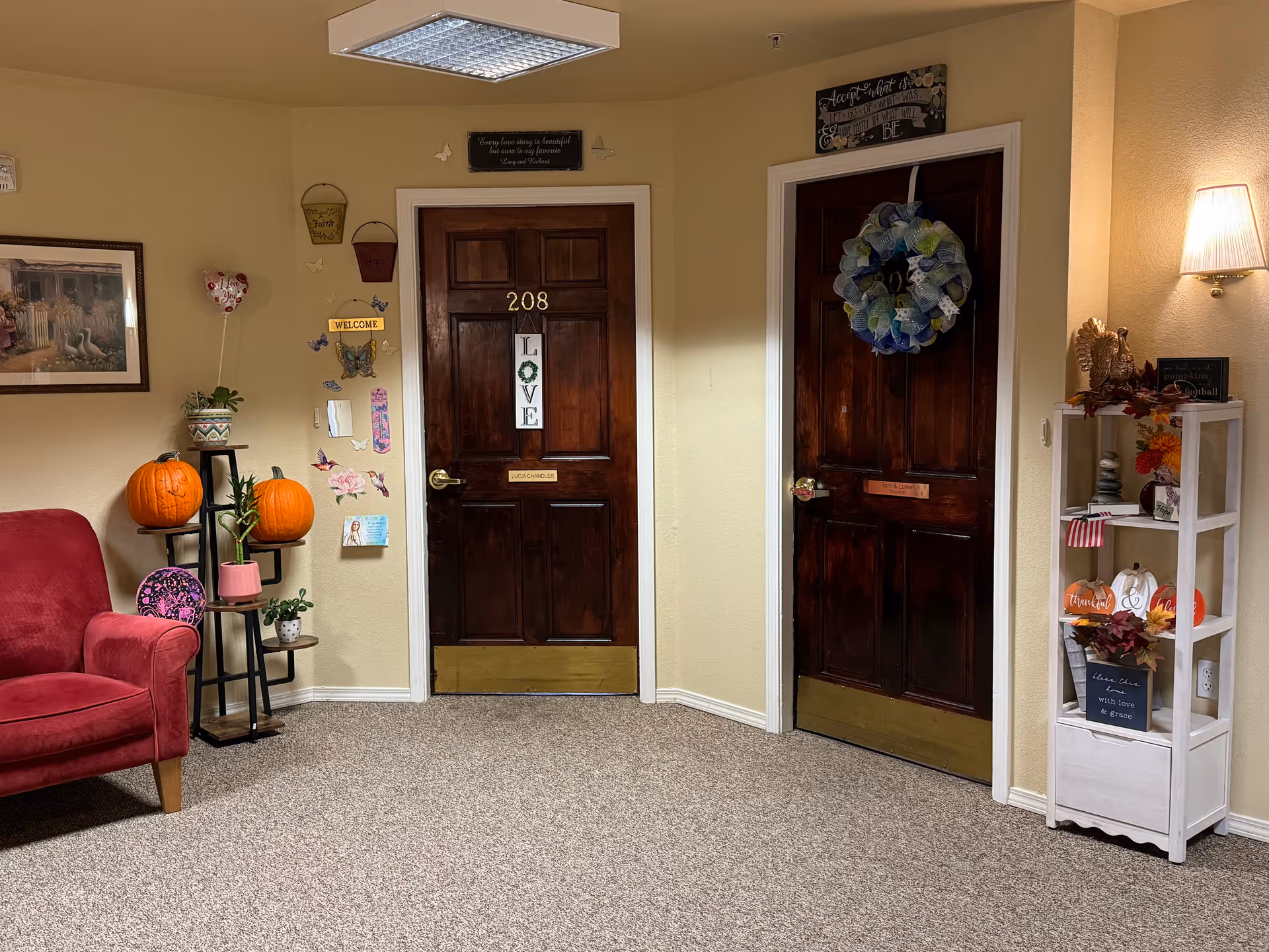 Interior hallway area with two dark wooden doors labeled 208 and another with a wreath. The walls are decorated with small signs, butterflies, and plants. There is a red armchair on the left and a white shelving unit with autumn decorations and a lamp on the right.
