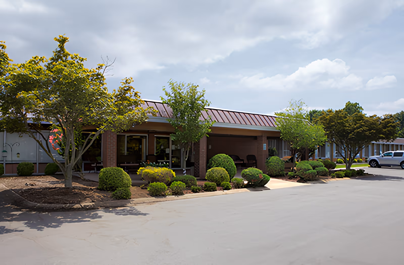 Exterior view of a single-story building with a covered entrance, surrounded by neatly trimmed bushes and trees under a partly cloudy sky. A few parked vehicles are visible in the background.