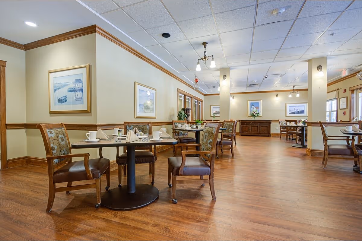A spacious dining room with wooden floors and multiple round tables set with white cups, plates, and folded napkins. The room features wooden chairs with cushioned seats and floral upholstery, beige walls with wooden trim, framed artwork, and ceiling lights.