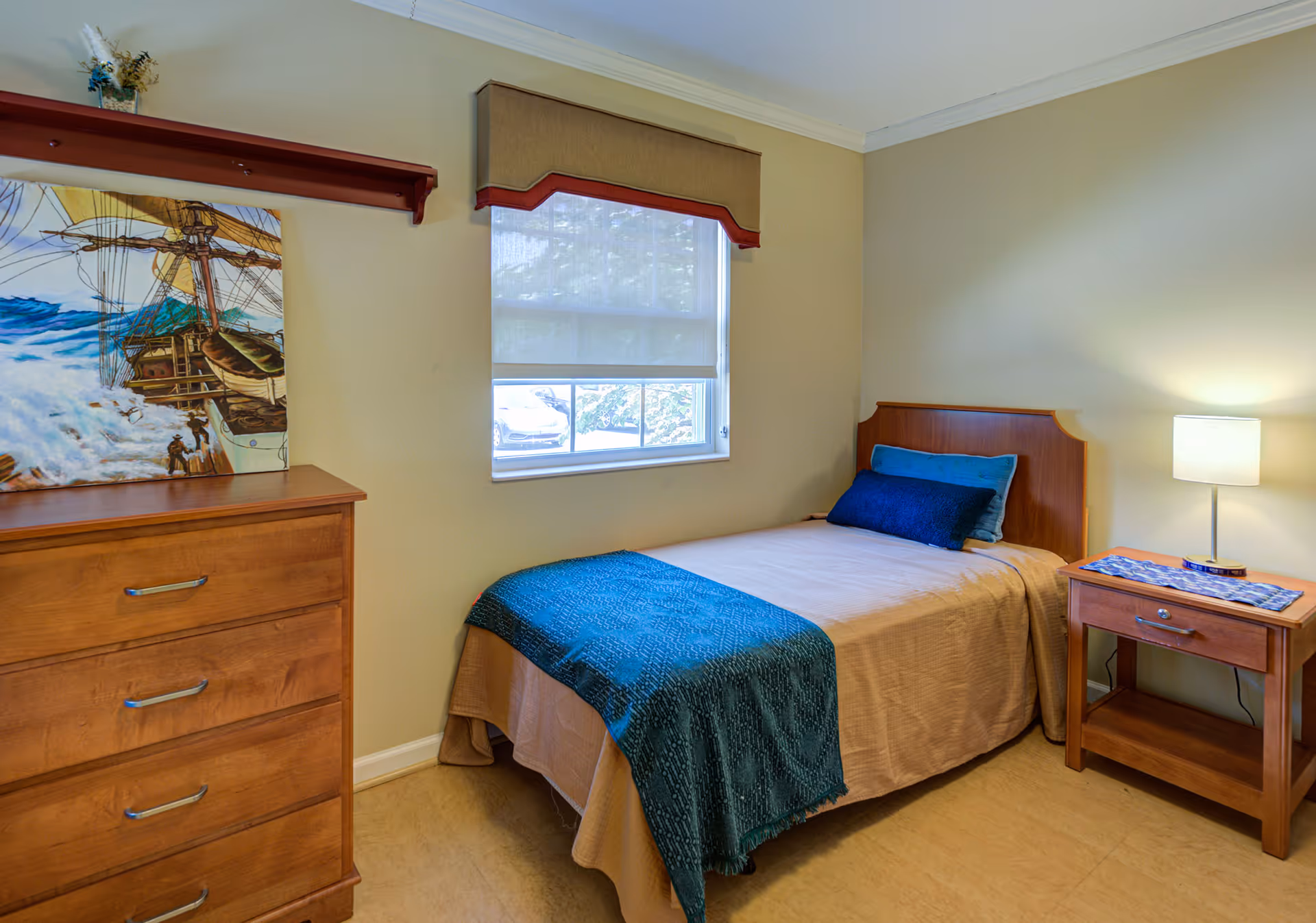 A small bedroom with a single bed covered in beige bedding and a blue throw blanket. There are two pillows on the bed, one blue and one dark blue. Next to the bed is a wooden nightstand with a lamp and a decorative cloth. On the left side of the room is a wooden dresser with four drawers and a painting of a ship resting on top. A window with a beige valance and a white roller shade is centered on the wall above the bed.