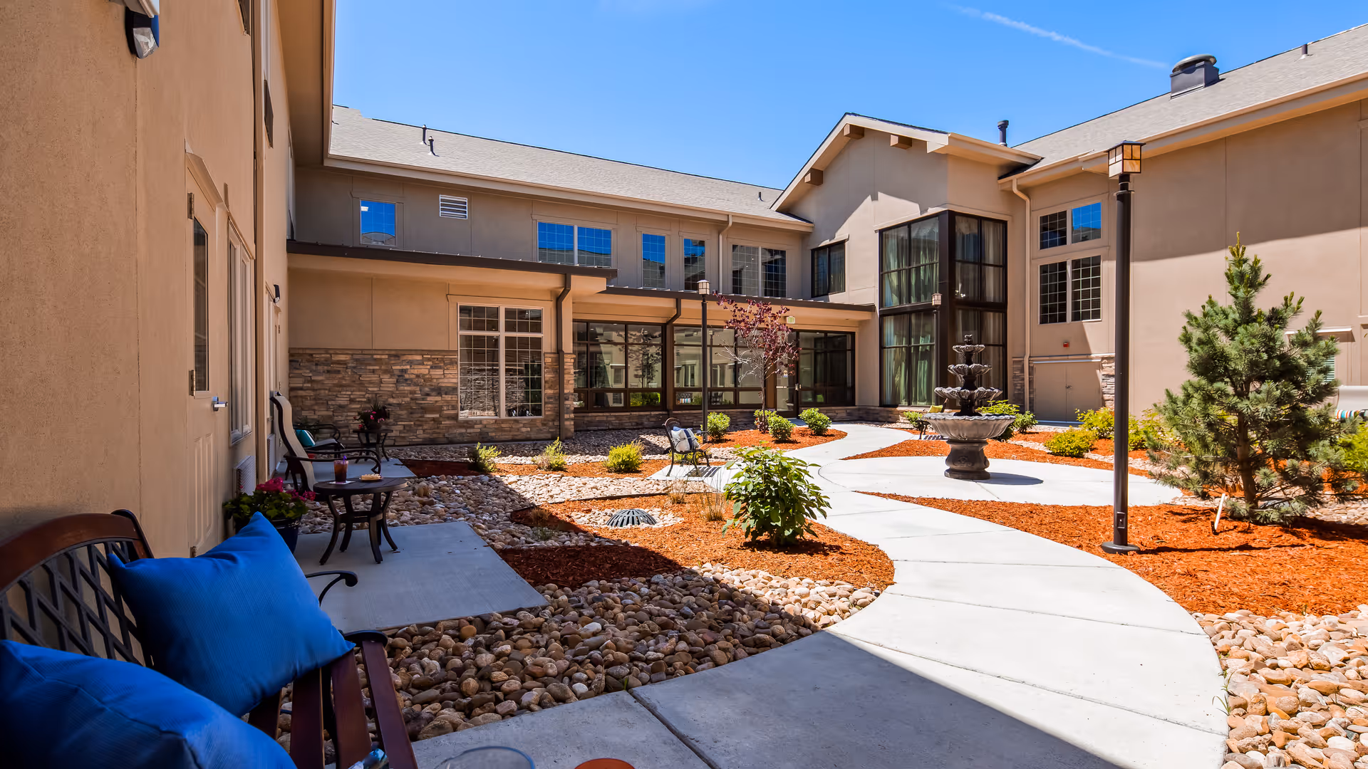 Outdoor courtyard area of a senior living facility with a curved concrete walkway, benches with blue cushions, small tables, a multi-tiered water fountain, landscaped plants, and a clear blue sky.