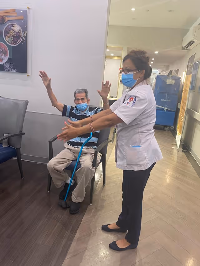 A healthcare worker wearing a white uniform and blue face mask is assisting an elderly man seated in a chair, who is also wearing a blue face mask and raising his arms. The setting appears to be an indoor facility with chairs and a hallway visible in the background.