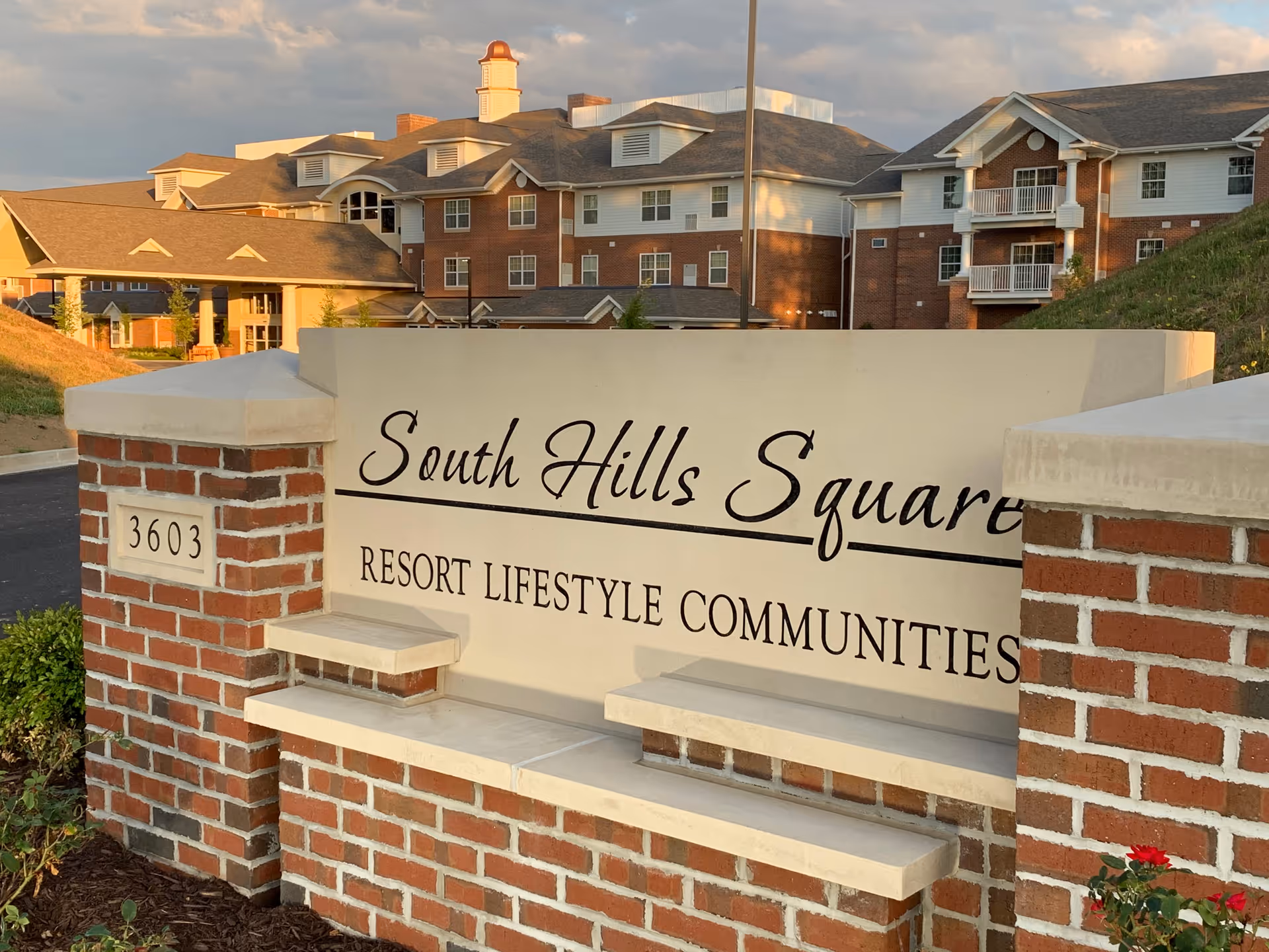 Entrance sign for South Hills Square Resort Lifestyle Communities with a large brick and stone sign in the foreground and a multi-story residential building in the background under a partly cloudy sky.