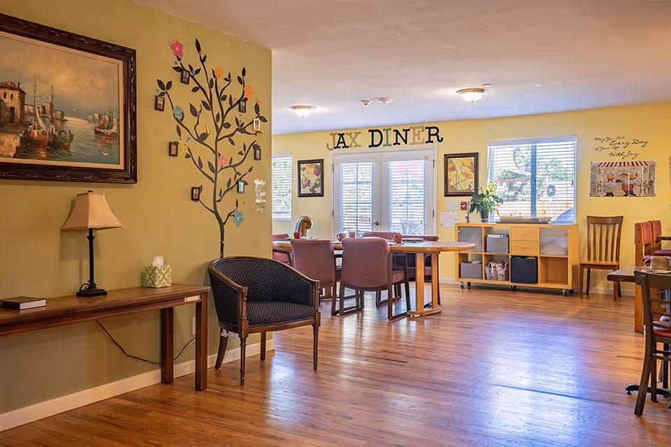 A bright and cozy dining area in an assisted living facility with wooden floors and yellow walls. There is a wooden table surrounded by chairs in the center, a decorative wall decal of a tree with colorful flowers, framed artwork, and a lamp on a wooden console table. The wall features the words 'JAX DINER' above double glass doors, and there are windows letting in natural light.