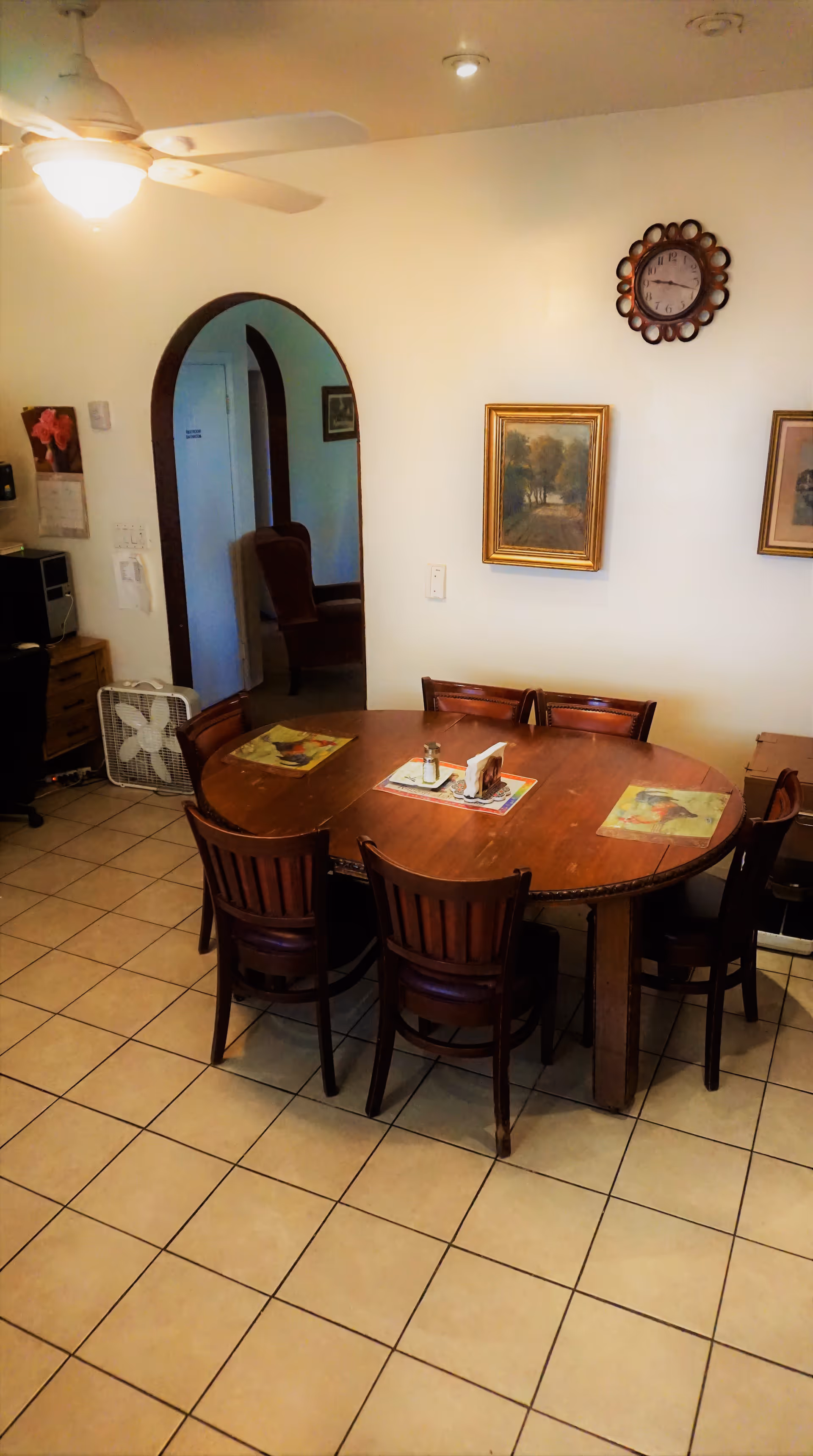 Wooden dining table with six chairs in a tiled dining area under a ceiling fan, with framed pictures and a wall clock.