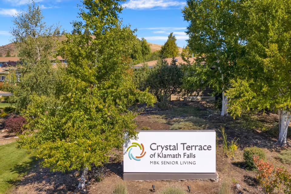 Outdoor view of the Crystal Terrace of Klamath Falls senior living facility sign surrounded by green trees and landscaping under a blue sky.