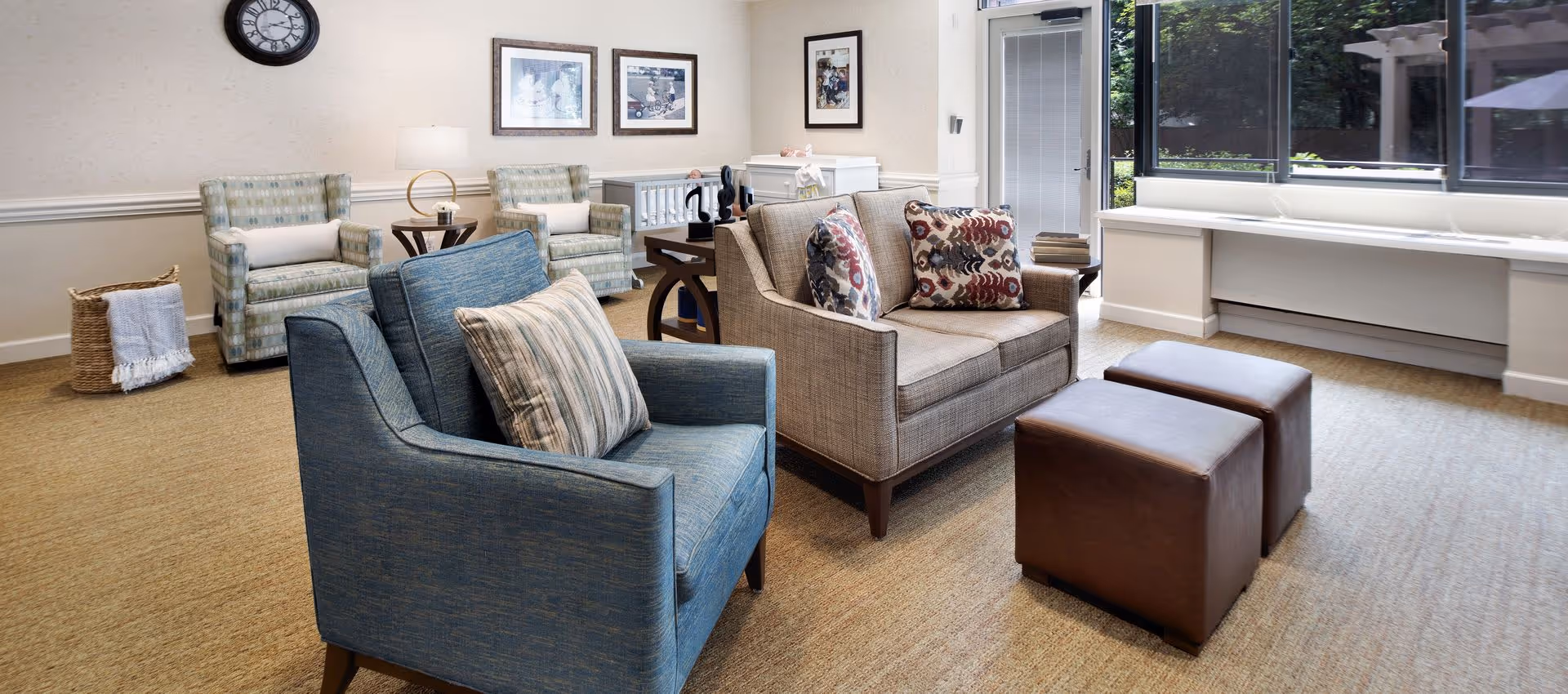 A bright communal living area with upholstered armchairs, a loveseat, two ottomans, wall art, and large windows overlooking greenery.
