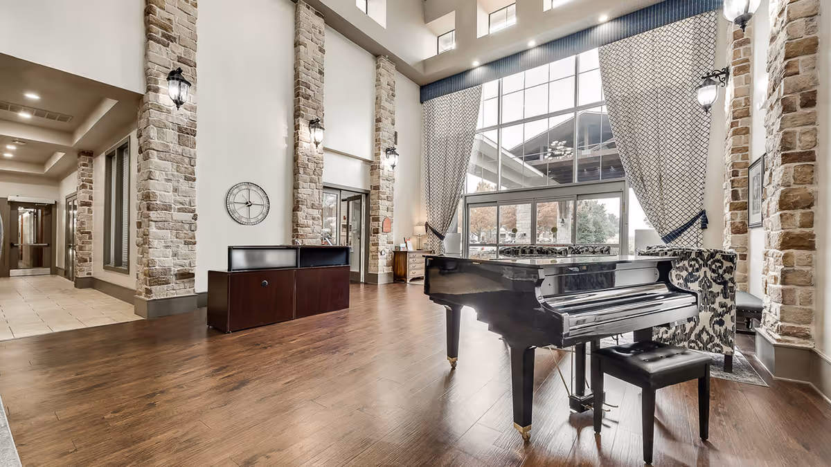 Spacious senior living lobby with a grand piano, tall windows, stone pillars, and a reception desk.