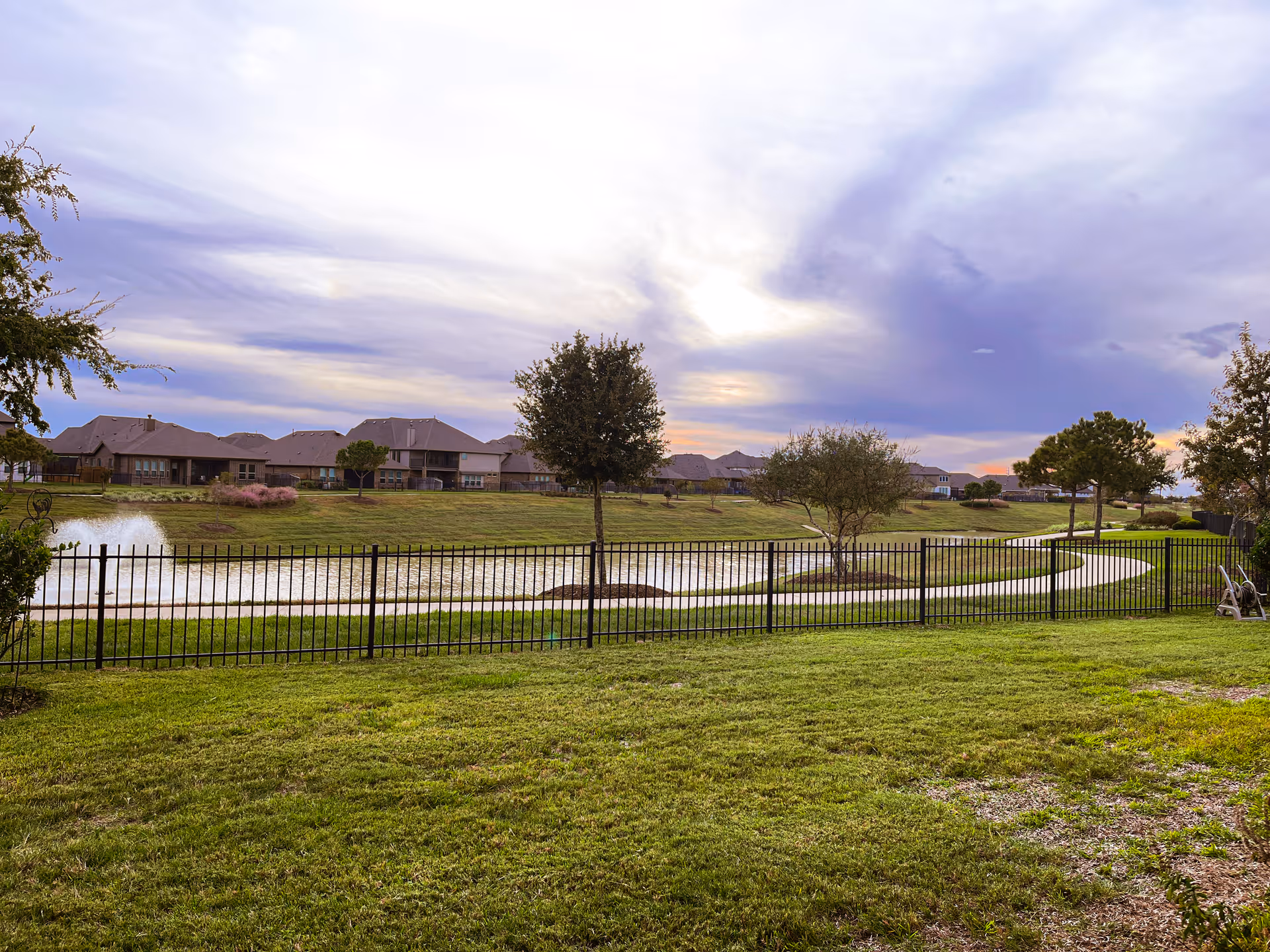 Grassy yard with a black metal fence overlooking a small pond and fountain with houses and trees under a cloudy sky.