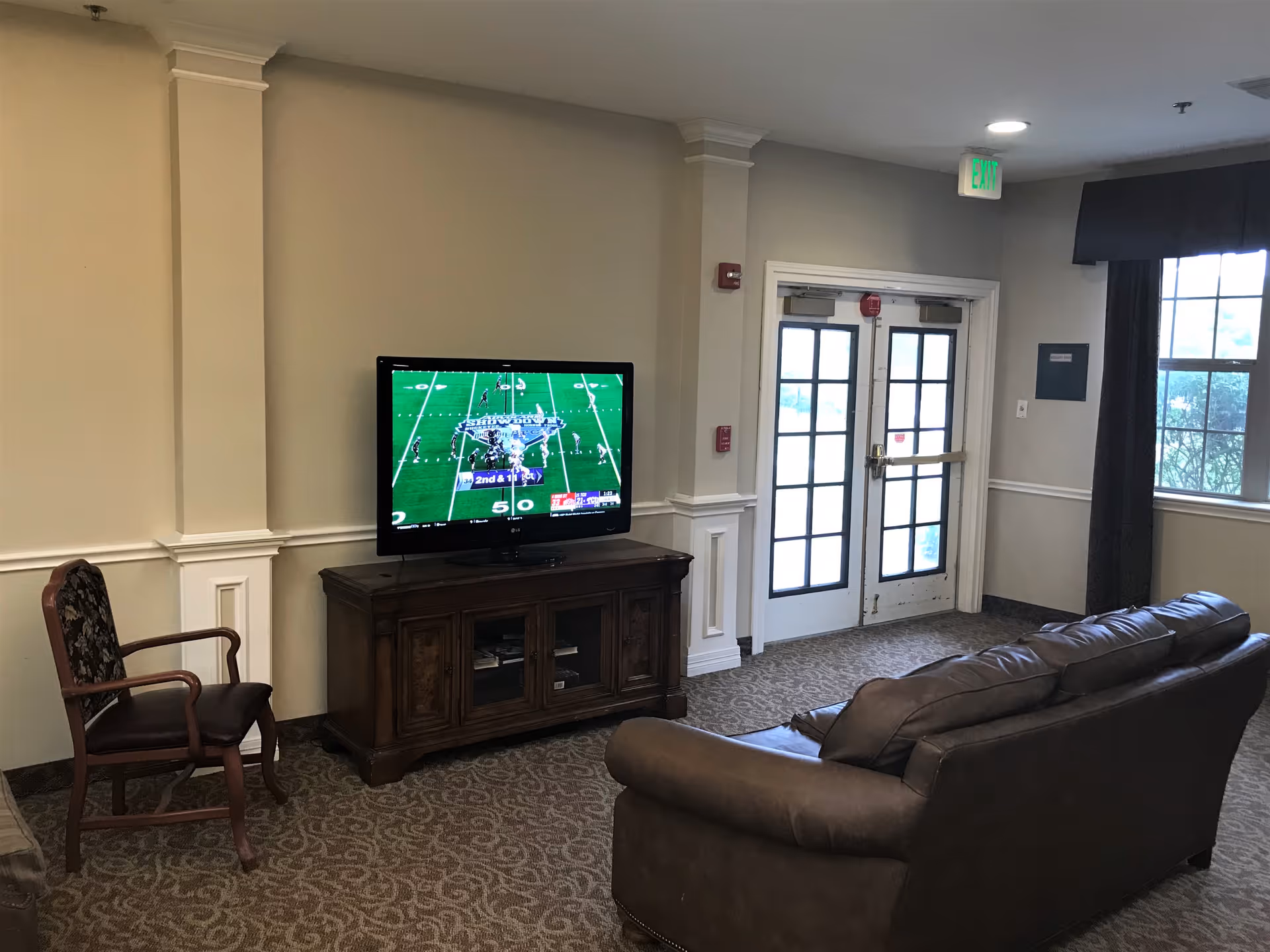 A cozy common area with a brown leather couch facing a flat-screen TV mounted on a wooden cabinet. The TV is displaying a football game. There is a wooden chair with a cushioned seat and backrest to the left of the TV. The room has beige walls, carpeted floor, a window with dark curtains, and a double glass door with an exit sign above it.