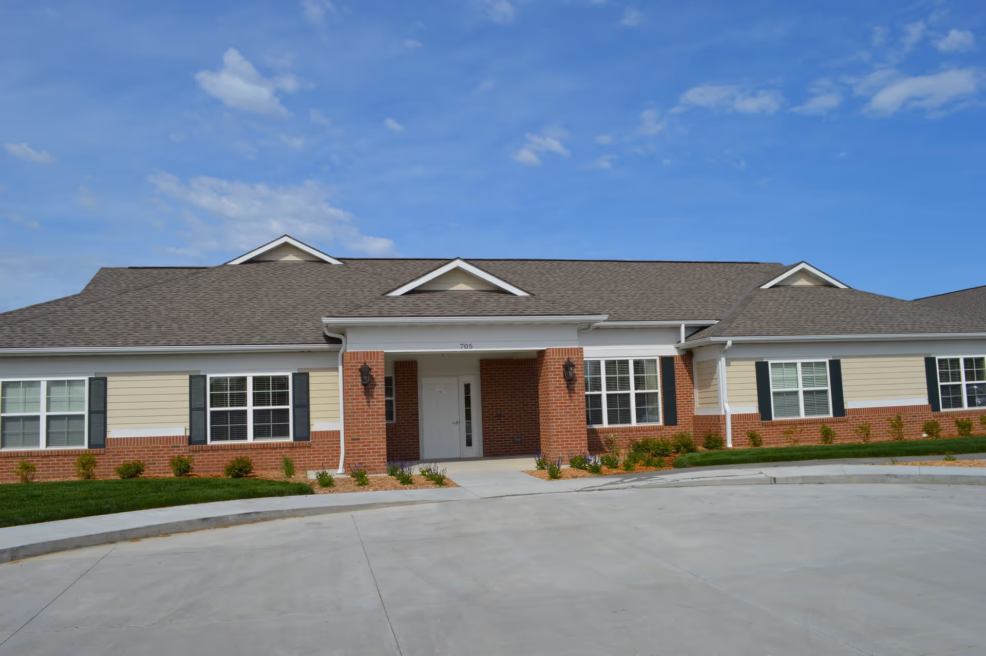 Front exterior view of a single-story building with beige siding and red brick accents, featuring a central entrance with a white door under a covered porch. The building has multiple windows with dark shutters and a gray shingled roof under a blue sky with scattered clouds.