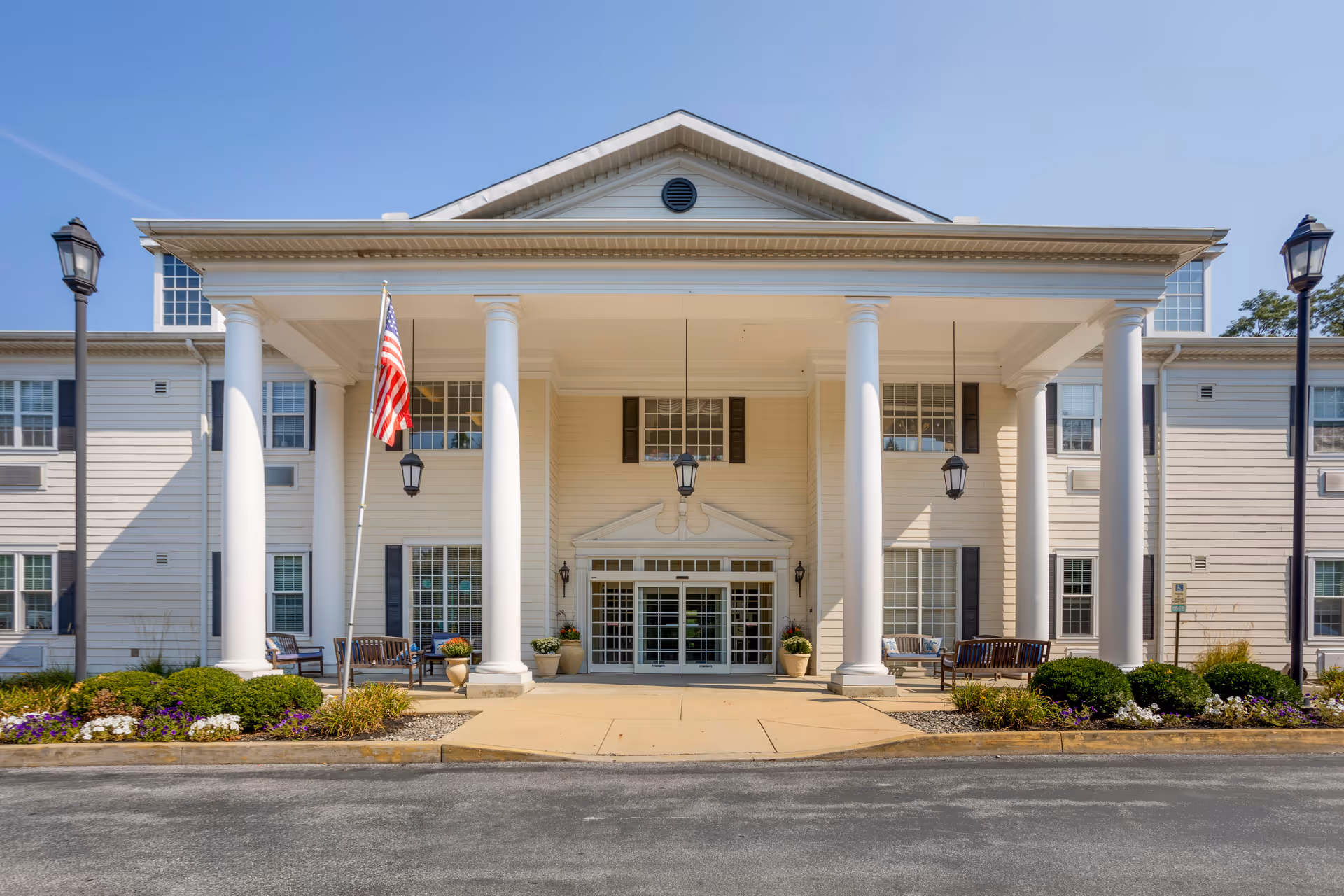 Front exterior view of a senior living facility with large white columns supporting a covered entrance. There are benches and flower pots on either side of the entrance, an American flag on a pole, and two street lamps. The building is light-colored with multiple windows and a clear blue sky above.