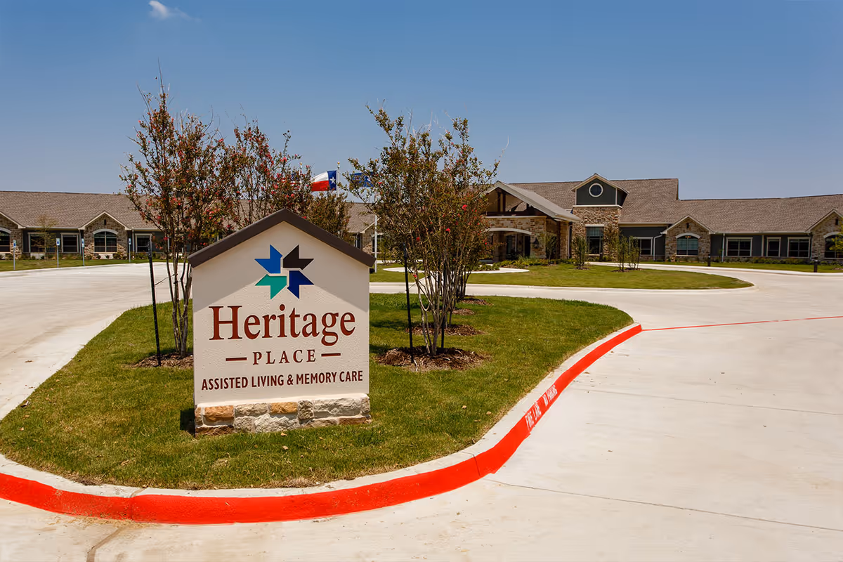 Exterior view of Heritage Place Assisted Living & Memory Care facility with a large sign in the foreground displaying the facility name and logo, surrounded by green grass and small trees, with a clear blue sky above.
