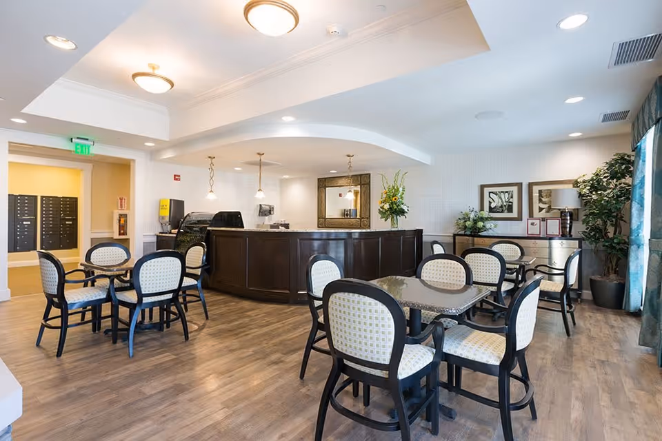 A bright and clean common area with several tables and chairs arranged for seating. The room features wood flooring, a dark wood reception desk or counter, pendant lights, a large mirror on the wall, and decorative plants. There is an exit sign above a doorway leading to a hallway with mailboxes.