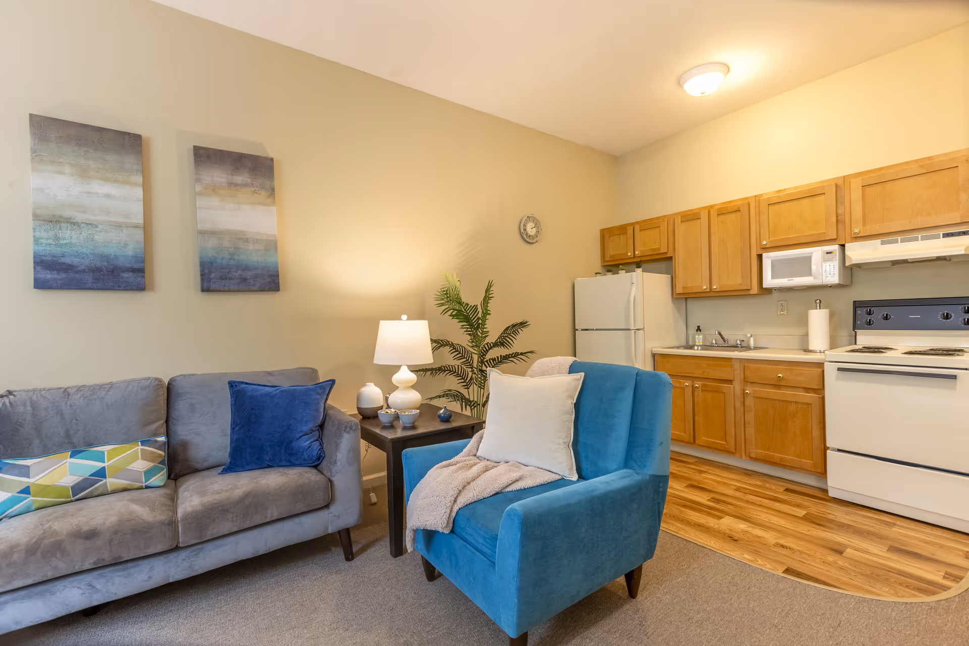 A cozy living room and kitchen area featuring a gray sofa with colorful pillows, a blue armchair with a white pillow and beige throw, a small dark wooden side table with a white lamp and decorative items, a potted plant, and a kitchen with wooden cabinets, white refrigerator, microwave, stove, and sink. The walls are light beige and the floor transitions from carpet in the living area to wood in the kitchen.