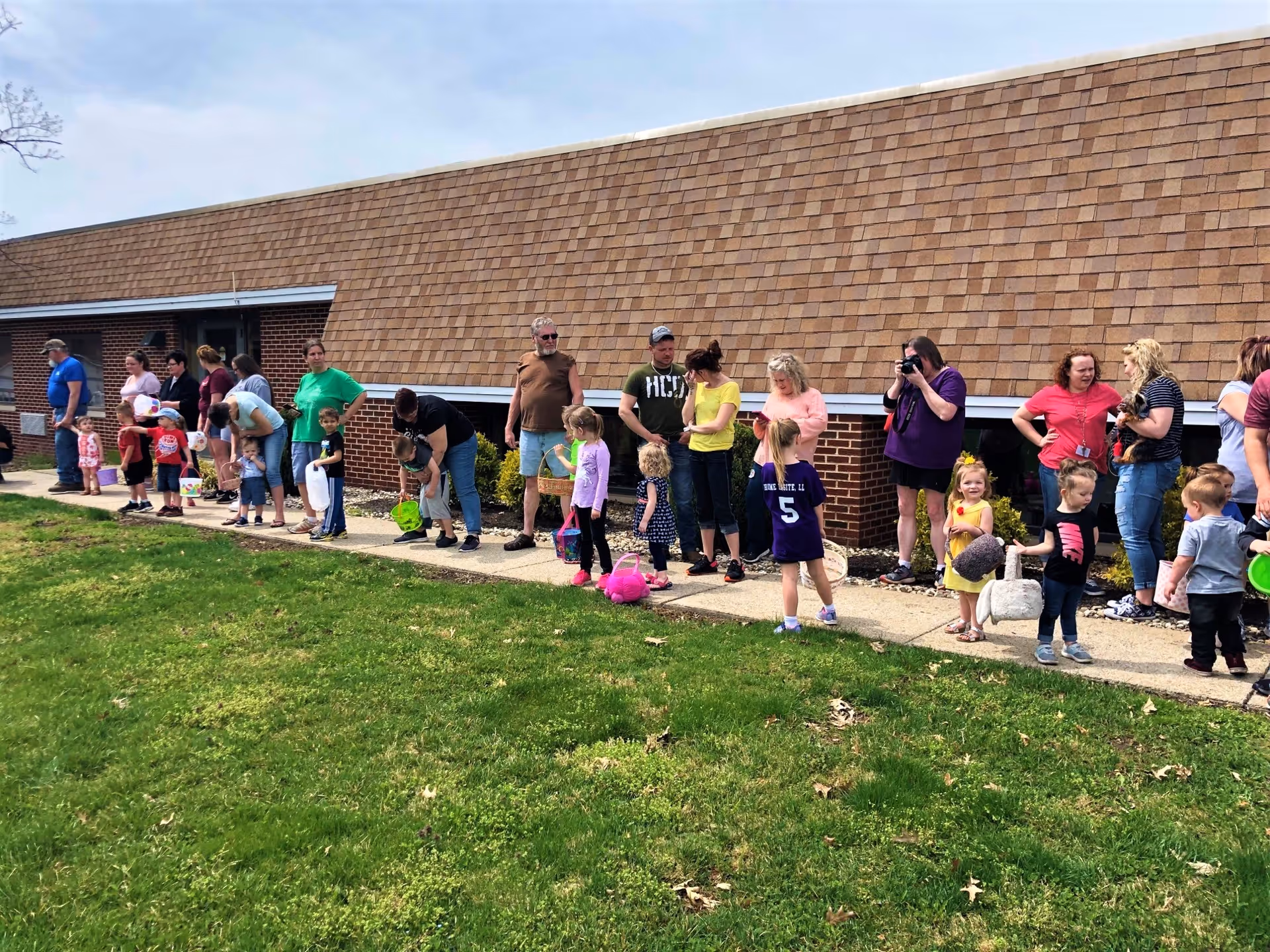 Children and adults lined up along a sidewalk outside a single-story brick nursing facility.