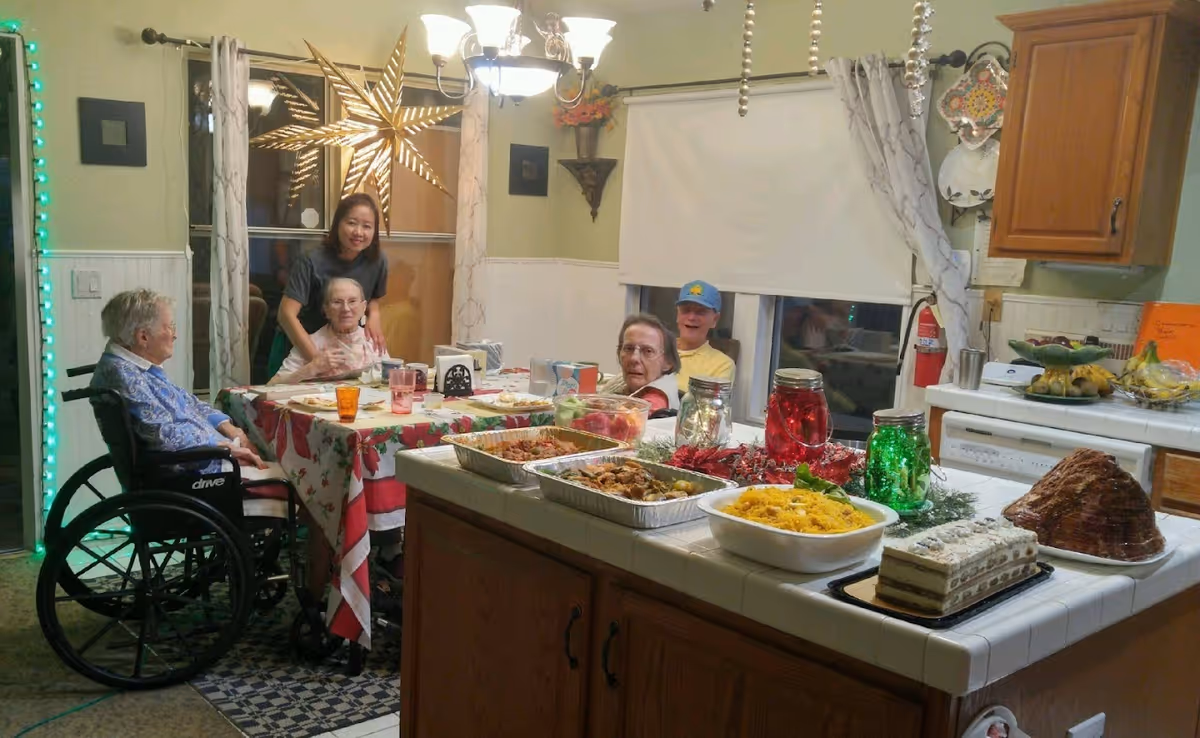 A group of elderly people and a caregiver gathered around a dining table in a cozy kitchen. The table is covered with a festive tablecloth and set with plates and cups. The kitchen counter nearby is filled with various dishes and desserts. The room is warmly lit with a chandelier and decorated with a large star hanging in the window and other decorative items.