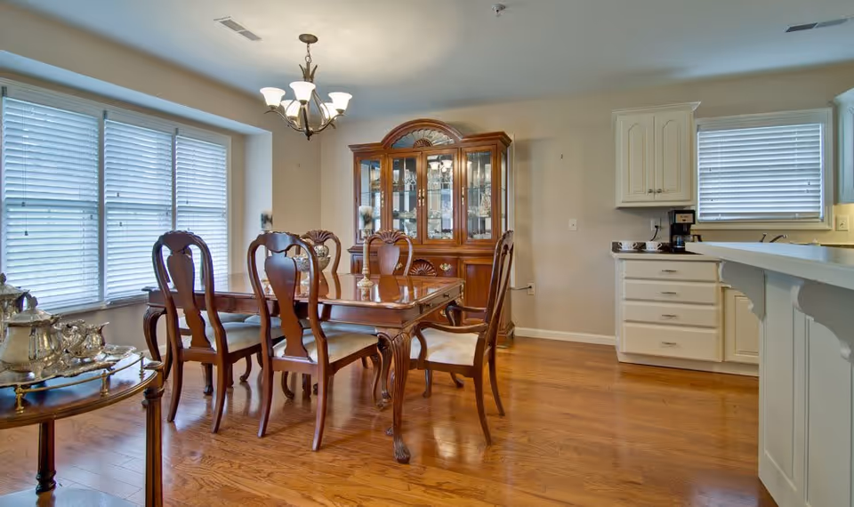 A dining room with a wooden dining table and six matching chairs with upholstered seats. There is a wooden china cabinet with glass doors against the wall. To the right, part of a kitchen with white cabinets, a coffee maker, and a window with blinds is visible. The floor is hardwood, and a chandelier hangs above the dining table. A small table with a silver tea set is on the left side near a large window with blinds.
