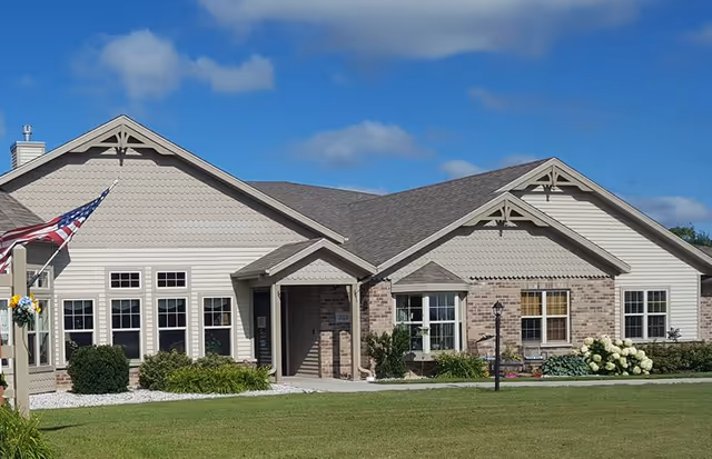 Exterior view of a single-story senior living facility building with beige siding and brick accents, multiple windows, a front entrance with a small covered porch, an American flag on a pole, and a well-maintained lawn with shrubs and flowers under a partly cloudy blue sky.
